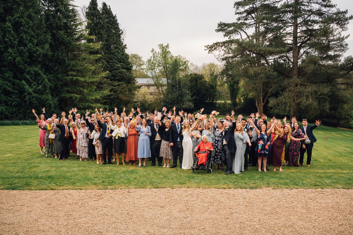 Large wedding party group photo on a lush green lawn with evergreen trees in the background.