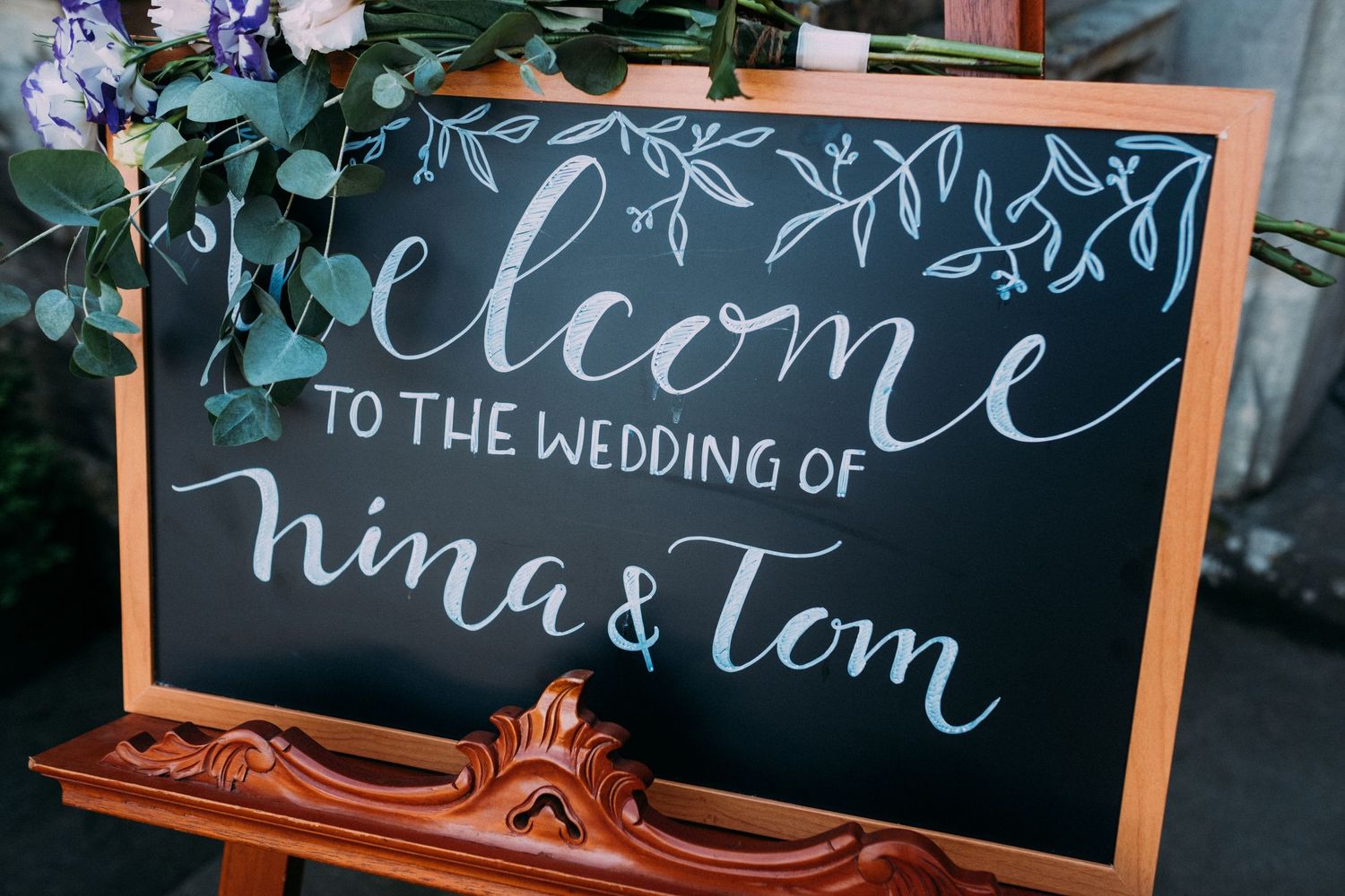 A wooden-framed chalkboard wedding welcome sign with elegant calligraphy announcing Nina and Tom's wedding.
