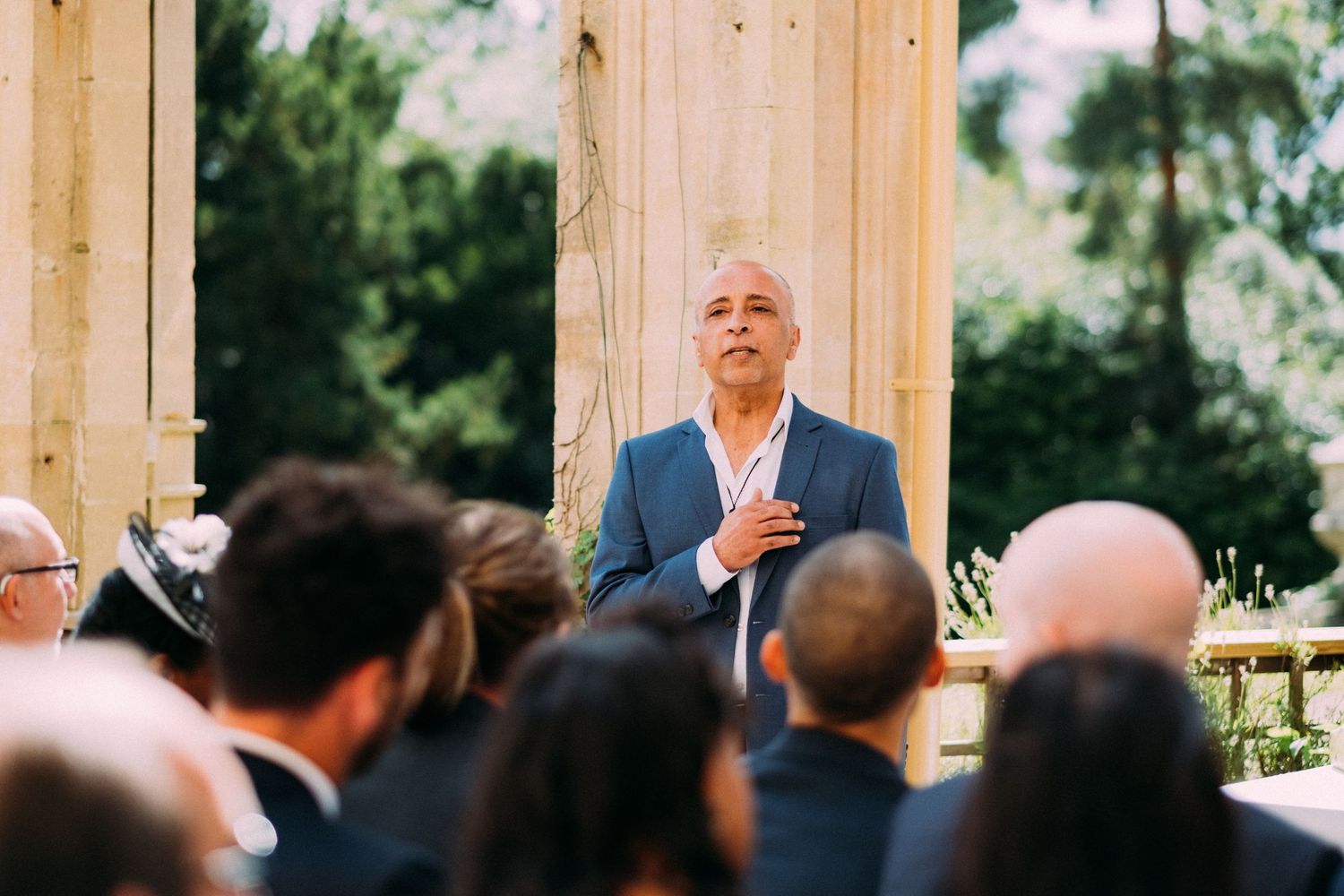 A speaker addresses wedding guests outdoors under classical columns during a ceremony.