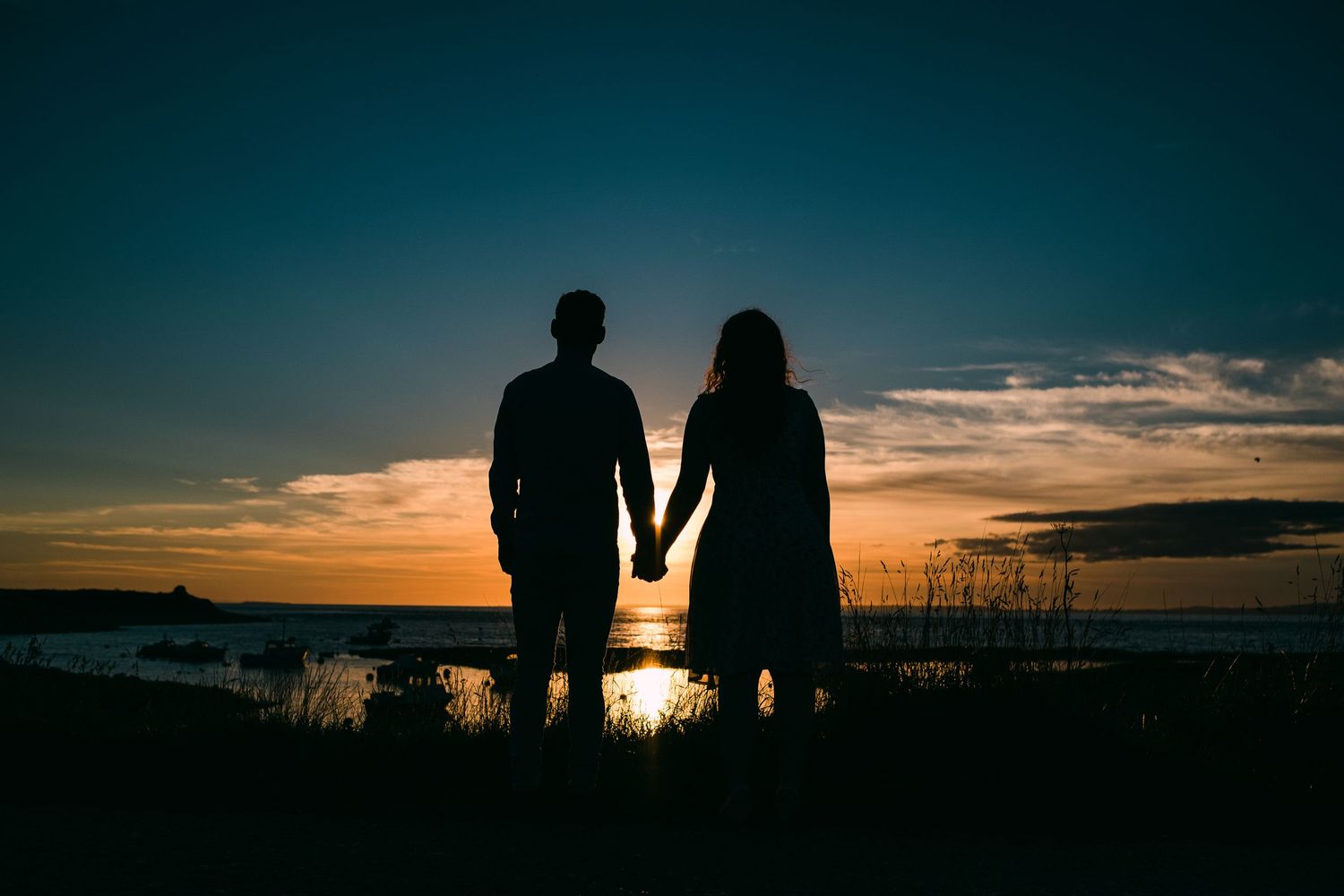 Silhouette of couple holding hands against dramatic sunset sky over water.