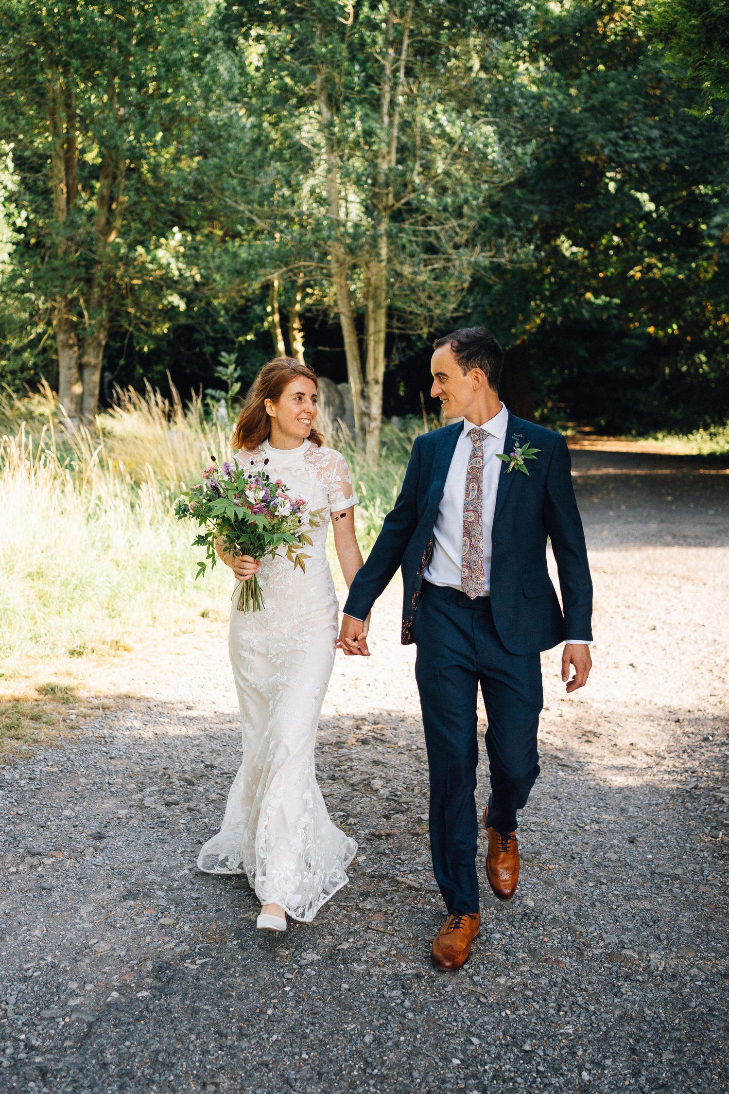 A couple walks and laughs together on a gravel path surrounded by trees during their wedding photoshoot.
