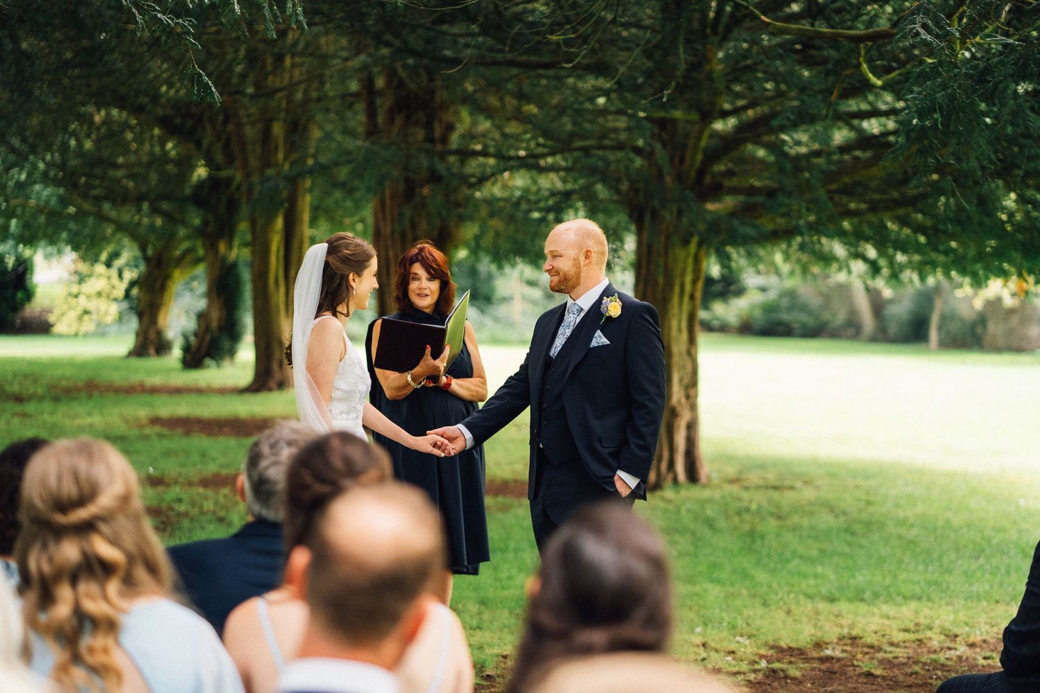 Garden wedding ceremony with guests seated on white chairs among trees.