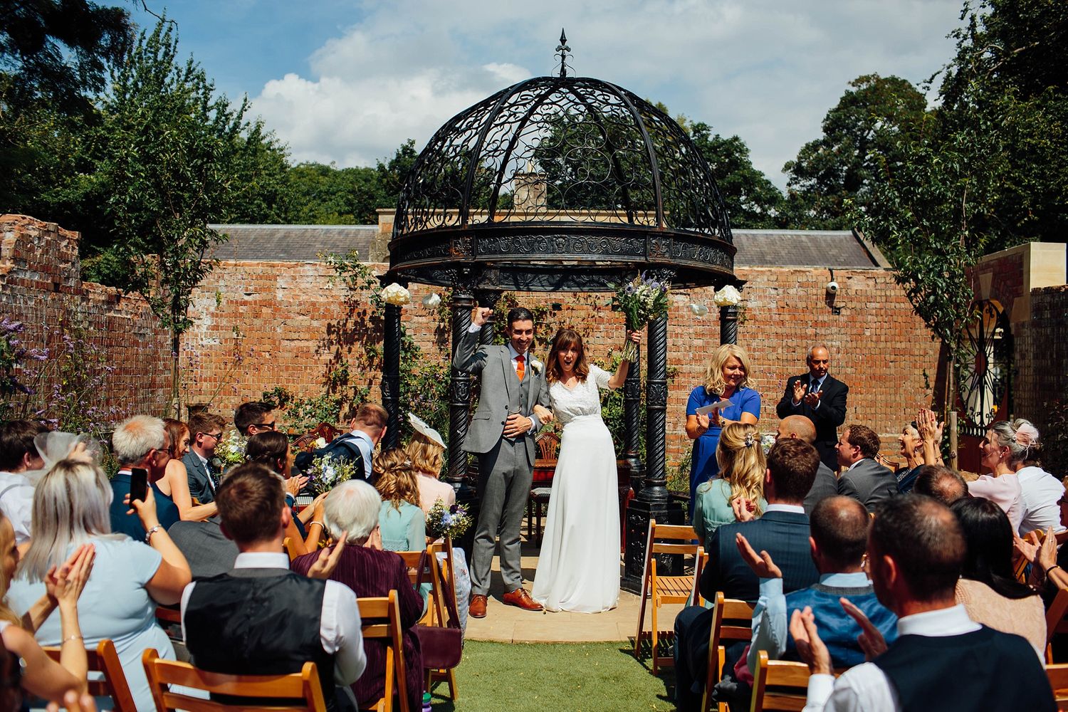 Panoramic view of outdoor wedding ceremony with gazebo and brick wall architecture.
