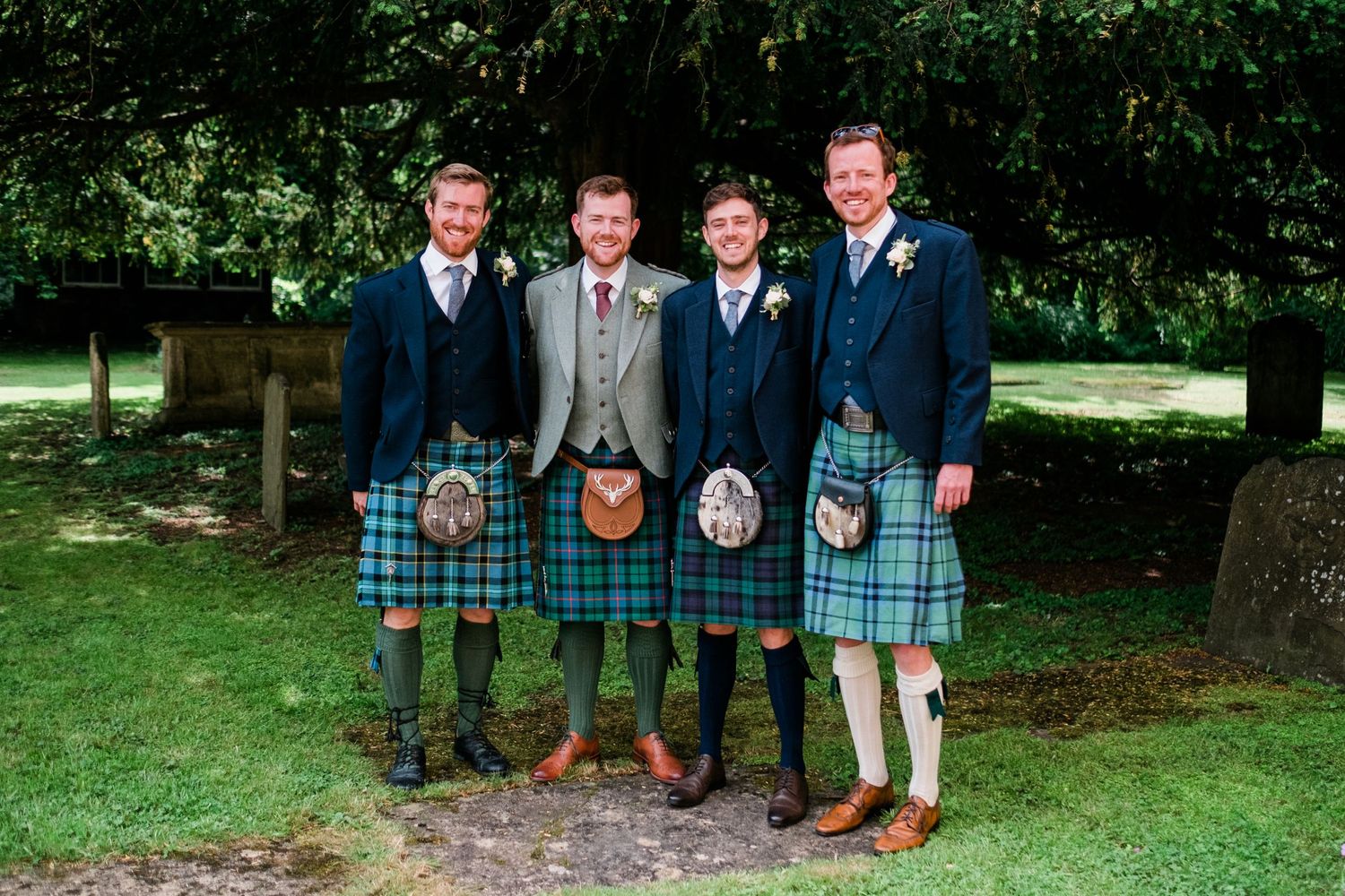 Groomsmen wearing traditional Scottish kilts pose together on grass near trees.