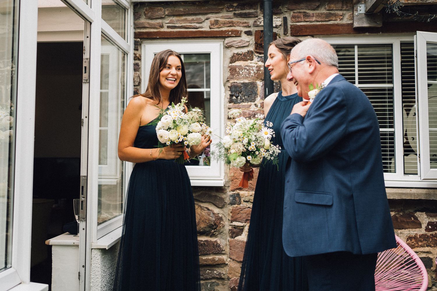 Wedding party members gather for candid reactions outside a rustic stone building.
