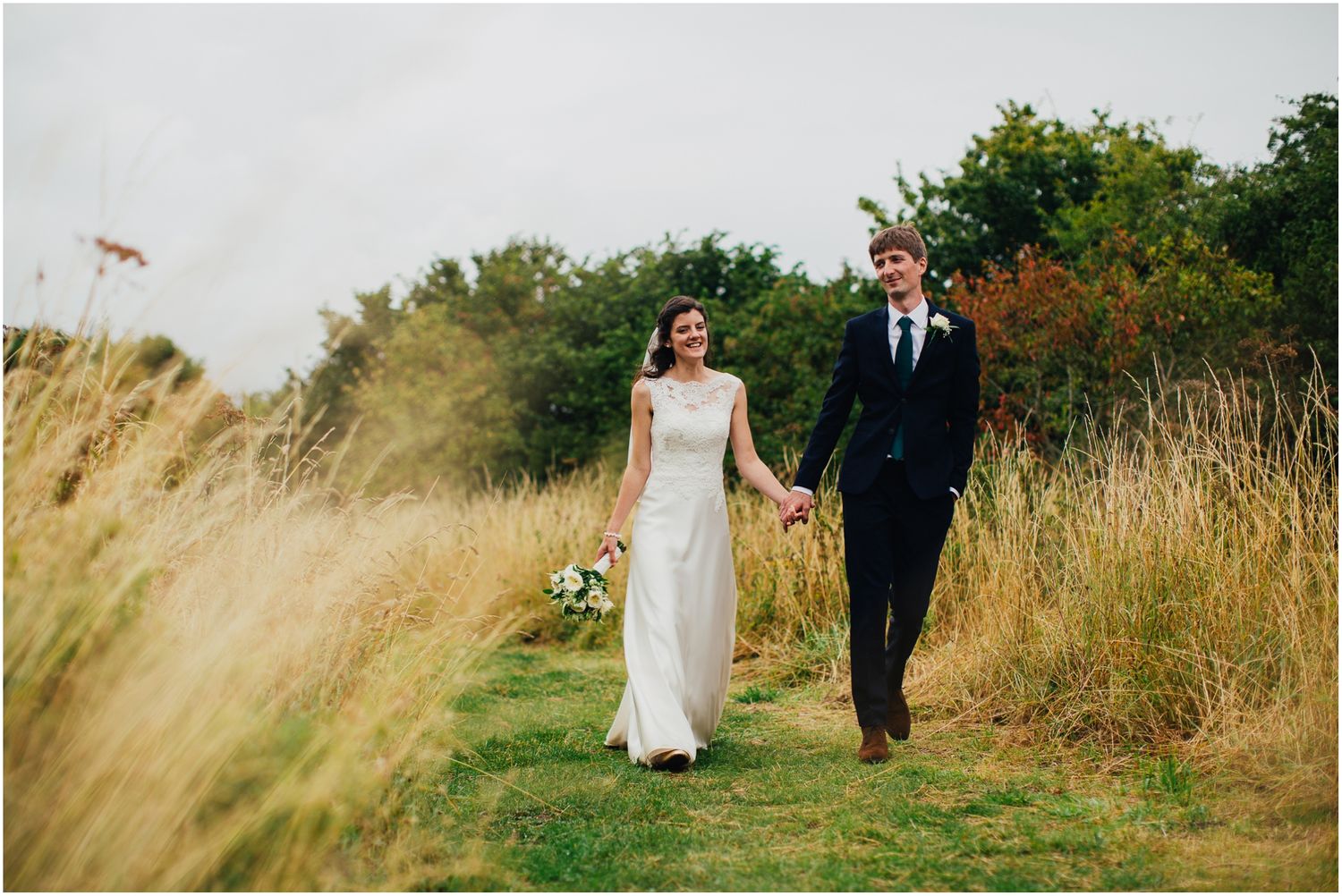Couple in wedding attire walks along path through natural meadow landscape.