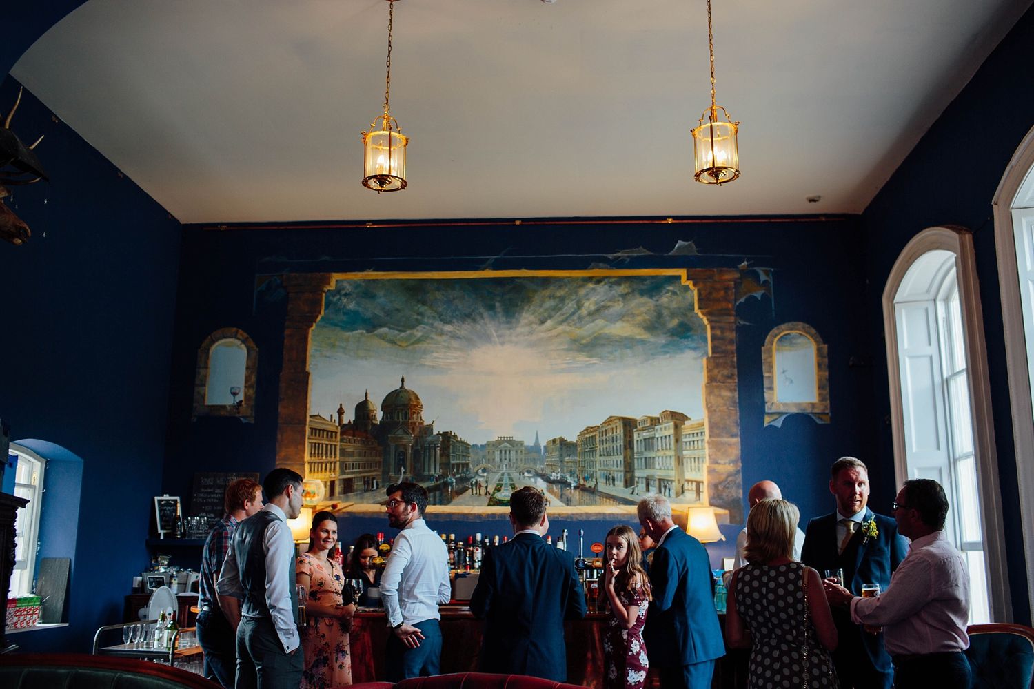 Guests gather in a blue room with hanging lanterns and a large cityscape mural during an event.