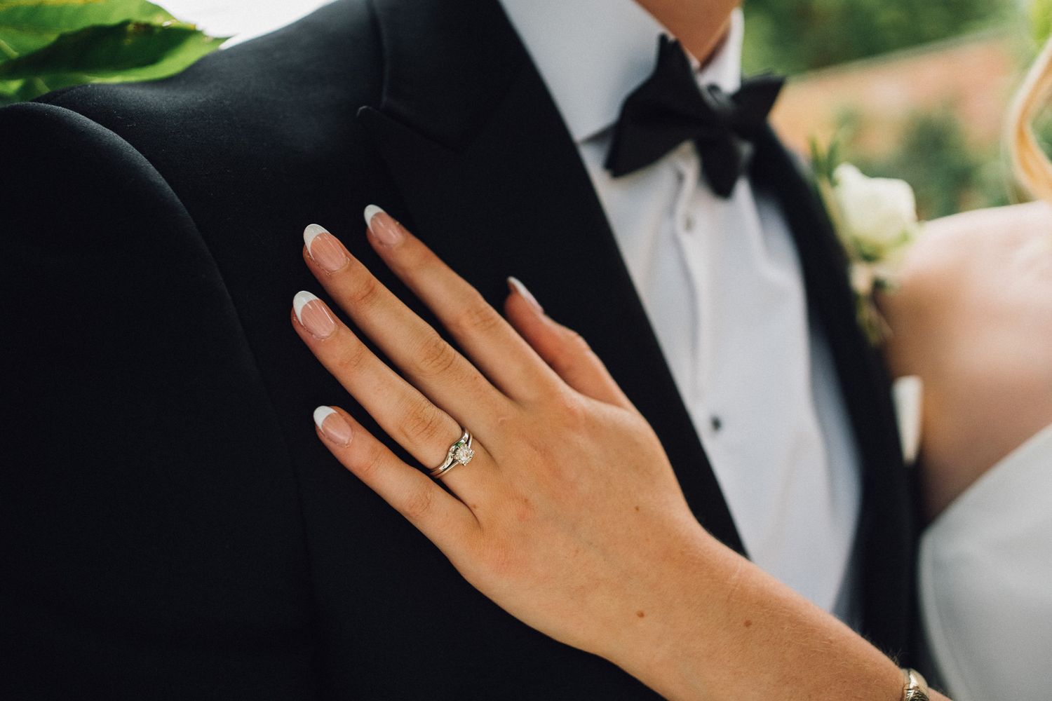 Close-up of an engagement ring on a hand with a formal tuxedo and white dress in the background.