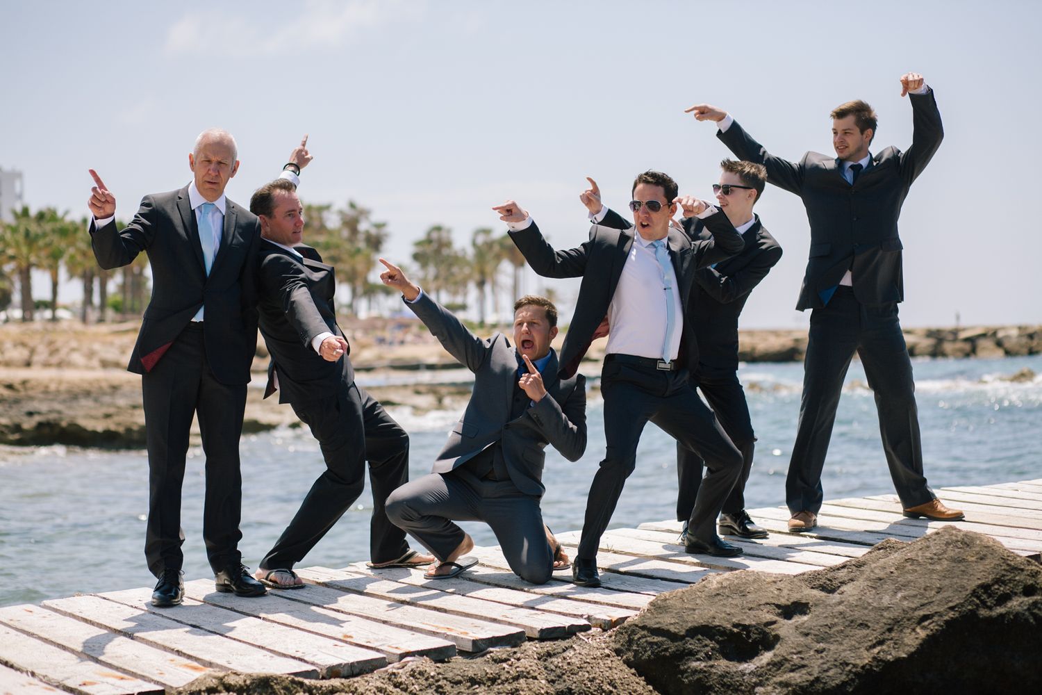 Wedding party members jump and pose energetically on rocks by the ocean.