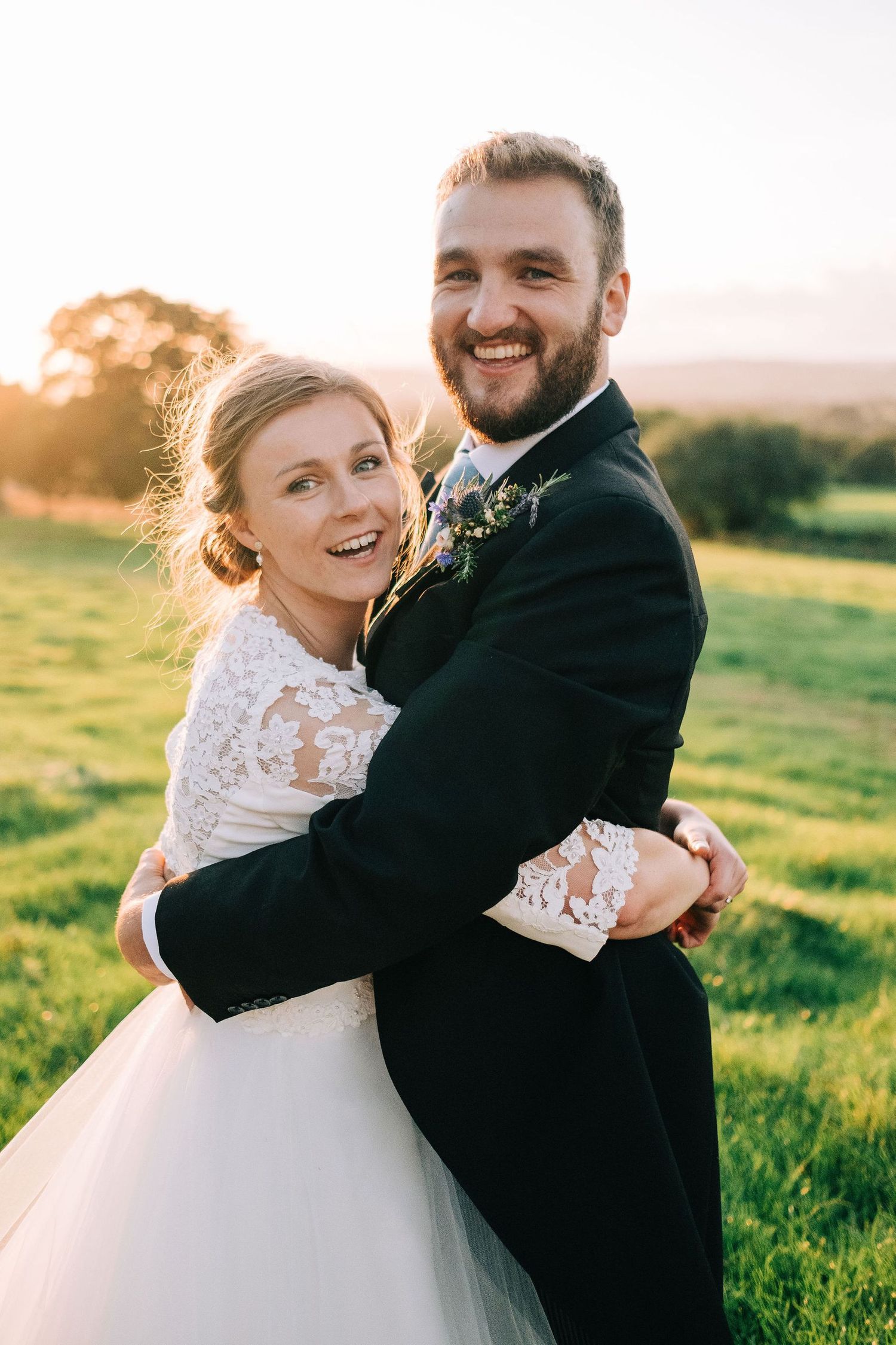 Wedding couple embraces and laughs together in a green field at sunset.