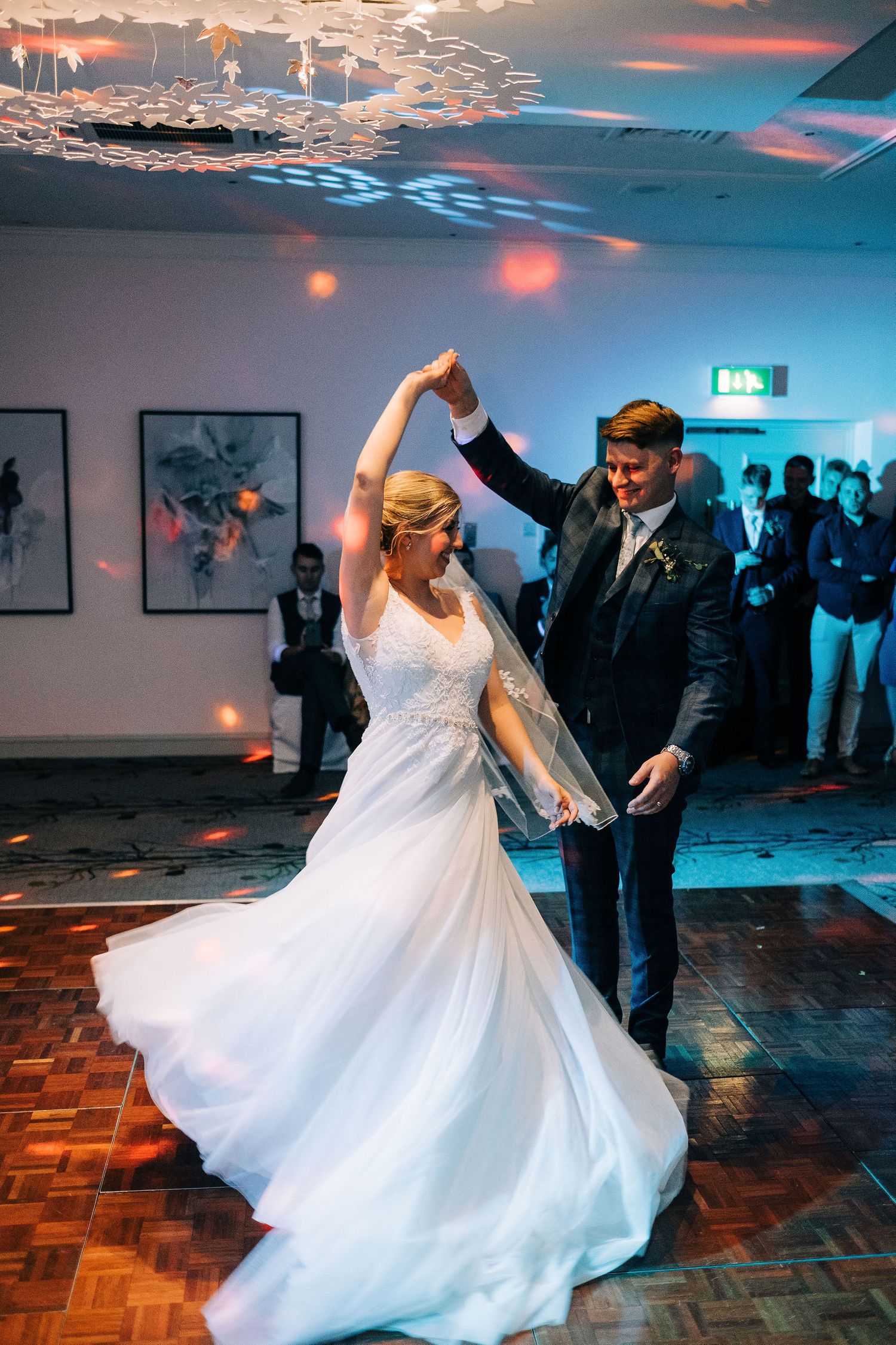 Bride in flowing white gown twirls during wedding dance under dramatic lighting.
