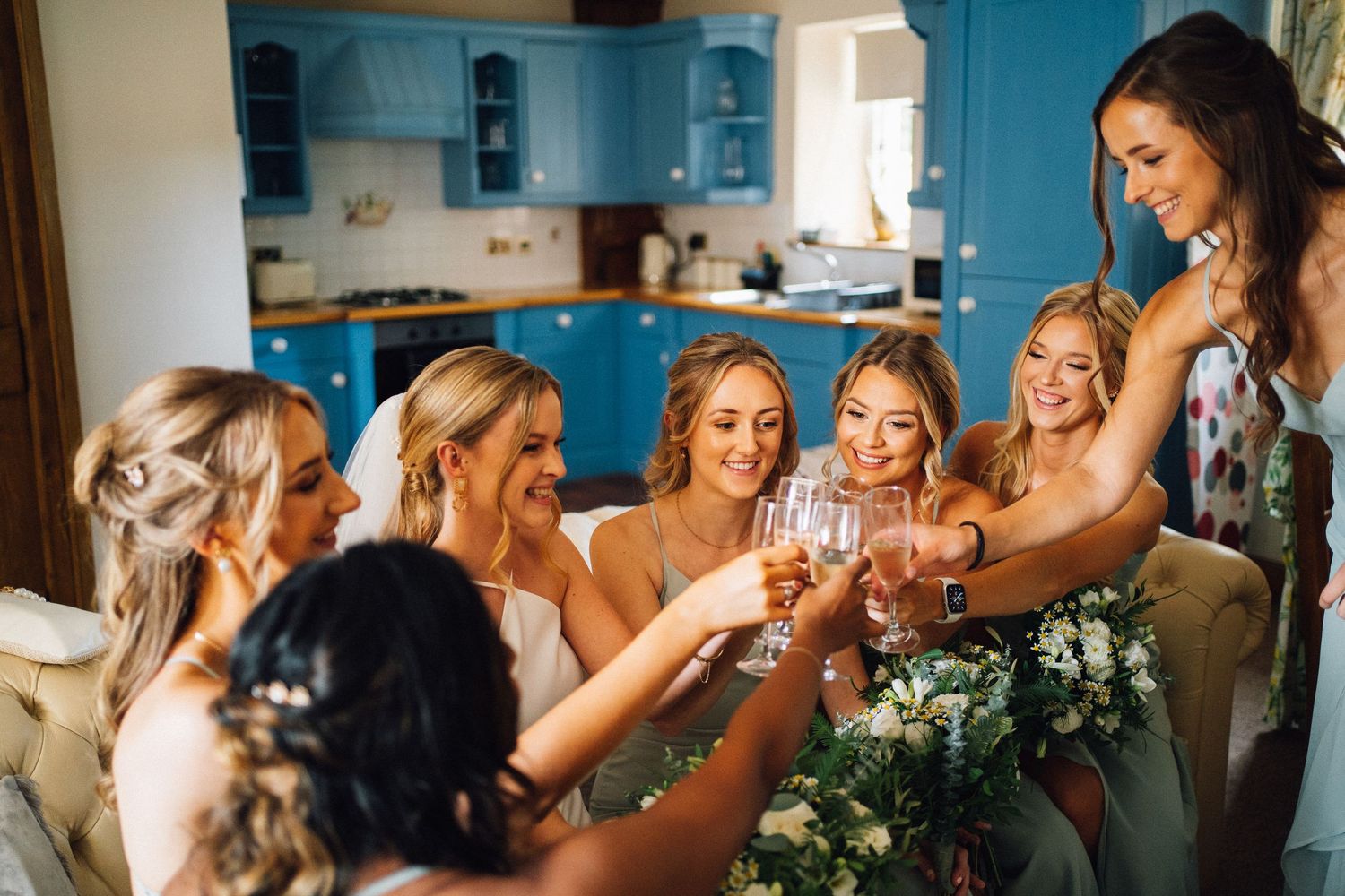 Bridesmaids in light blue dresses toast with champagne glasses in a kitchen with blue cabinets.