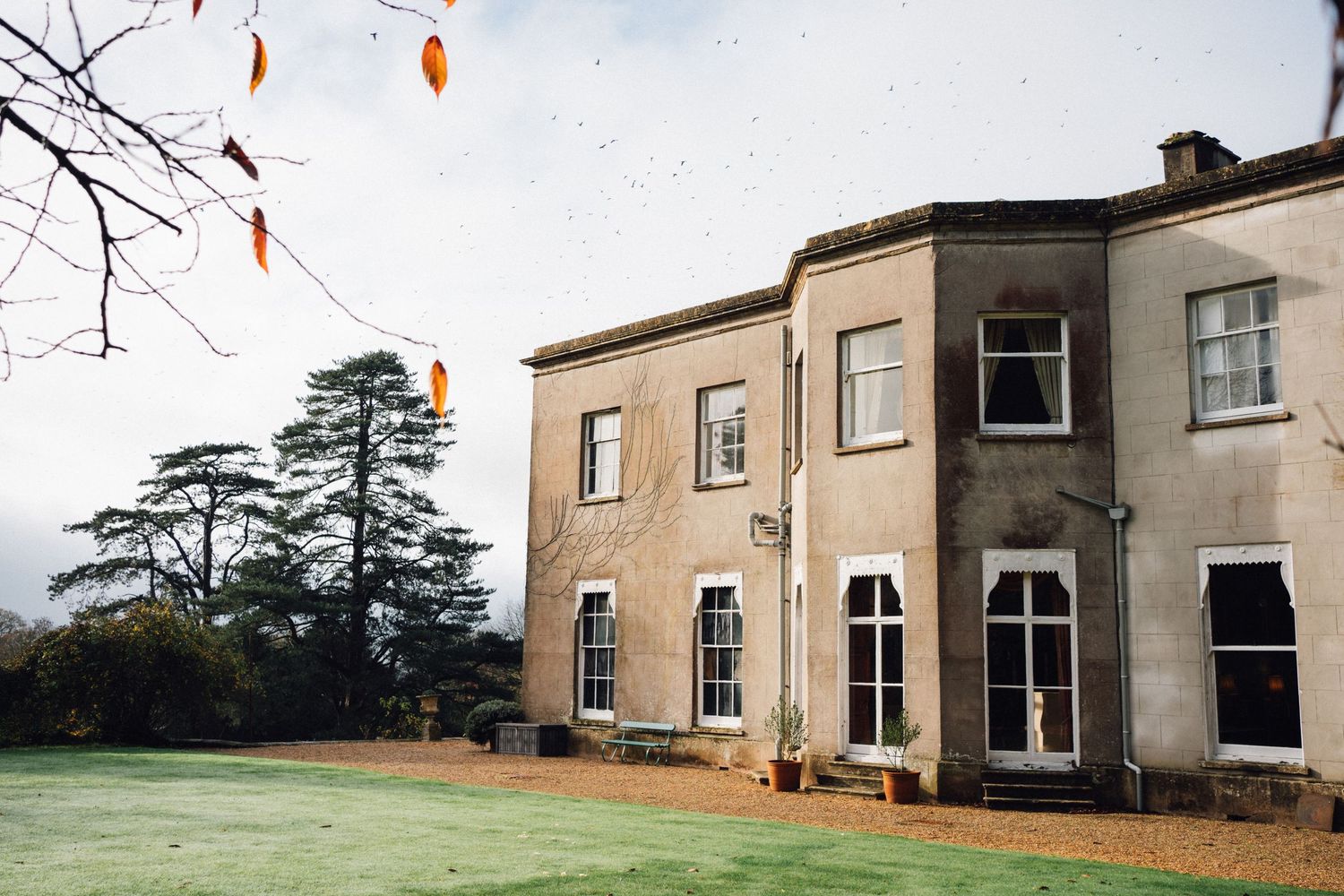 Stately beige stone manor house with arched windows and tall evergreen trees on a misty autumn lawn.