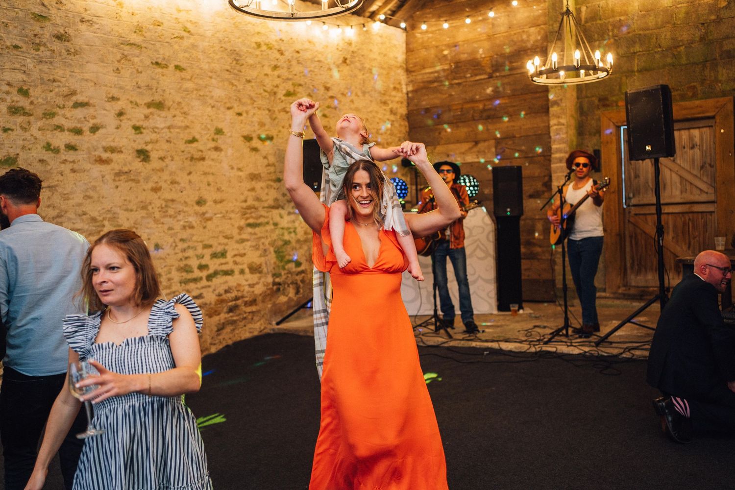 Wedding guest in bright orange dress dances enthusiastically on dark floor under warm lighting in stone venue.