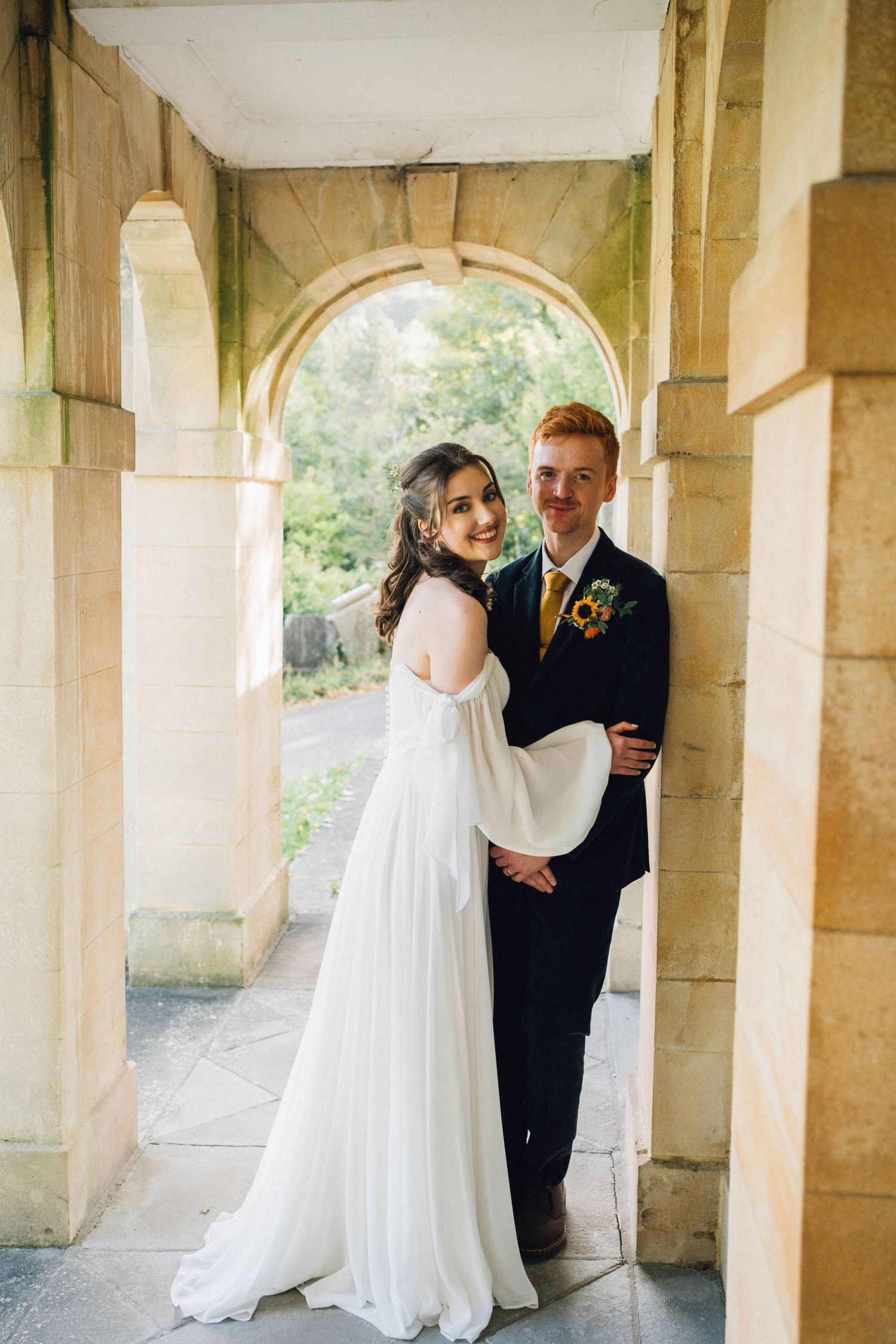A romantic wedding couple shares an intimate moment under a stone colonnade walkway.