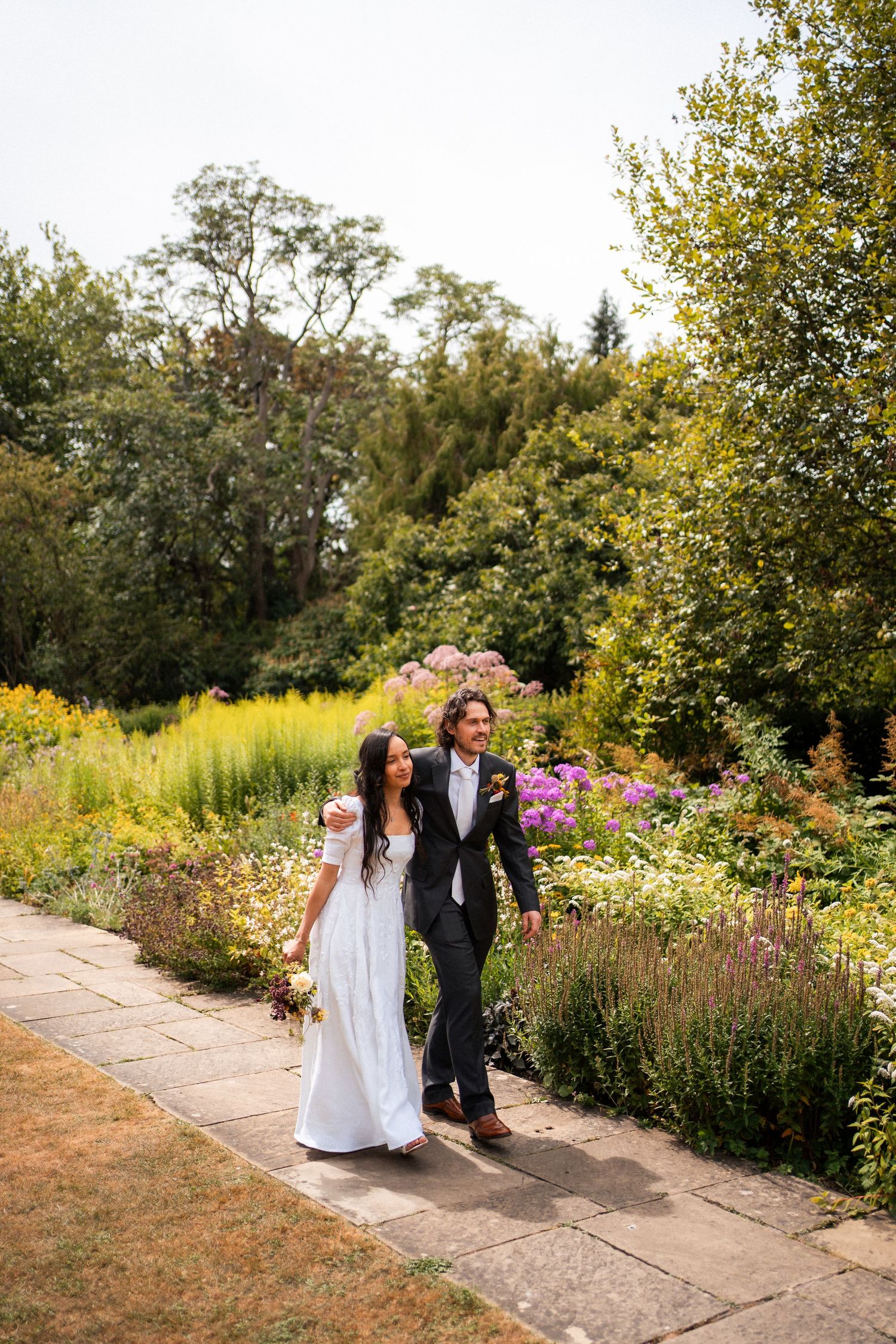 Bride and groom walking hand in hand along a garden path surrounded by colorful blooming flowers on their wedding day.