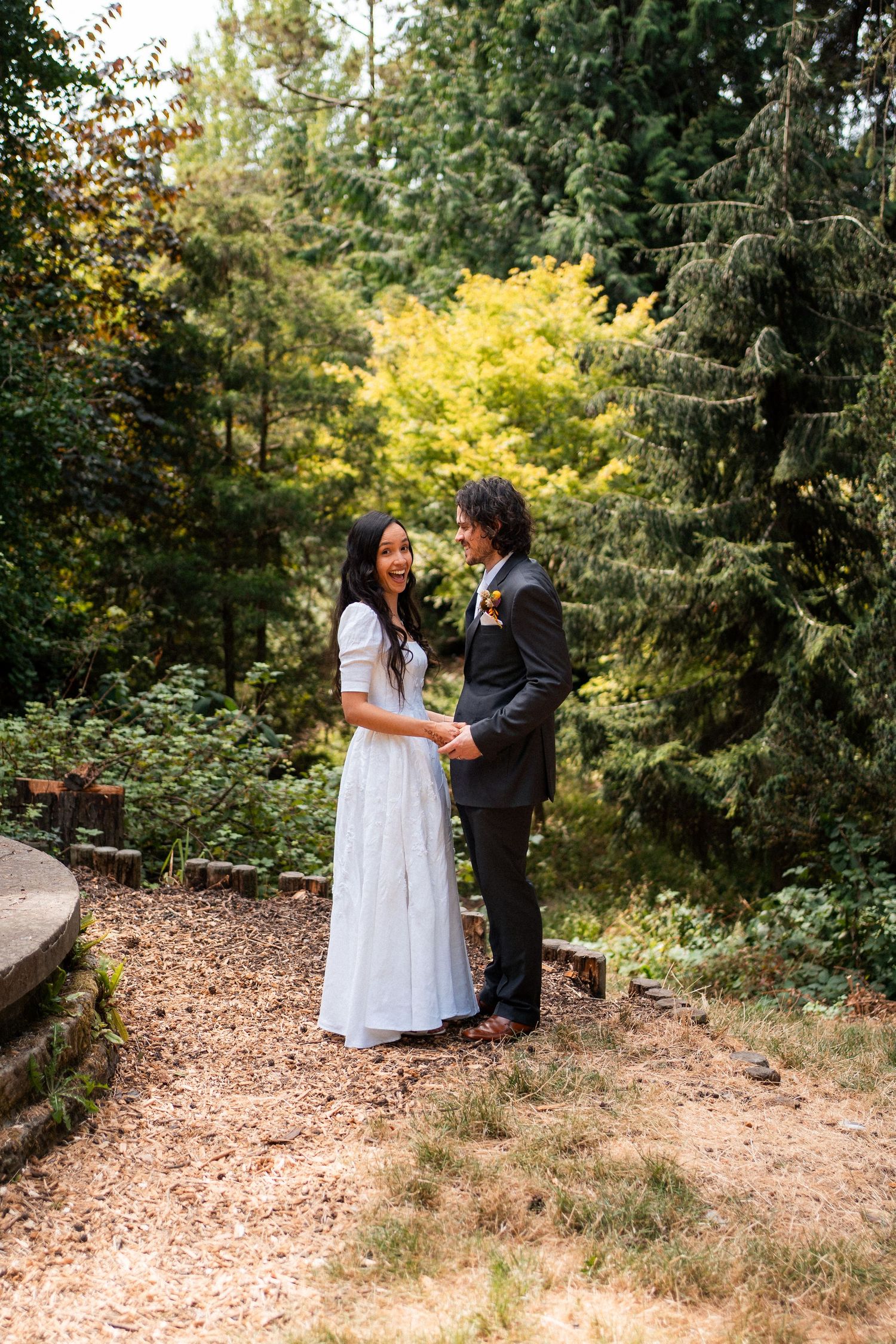 Bride and groom hold hands in a lush forest garden setting, bride wearing elegant white dress.
