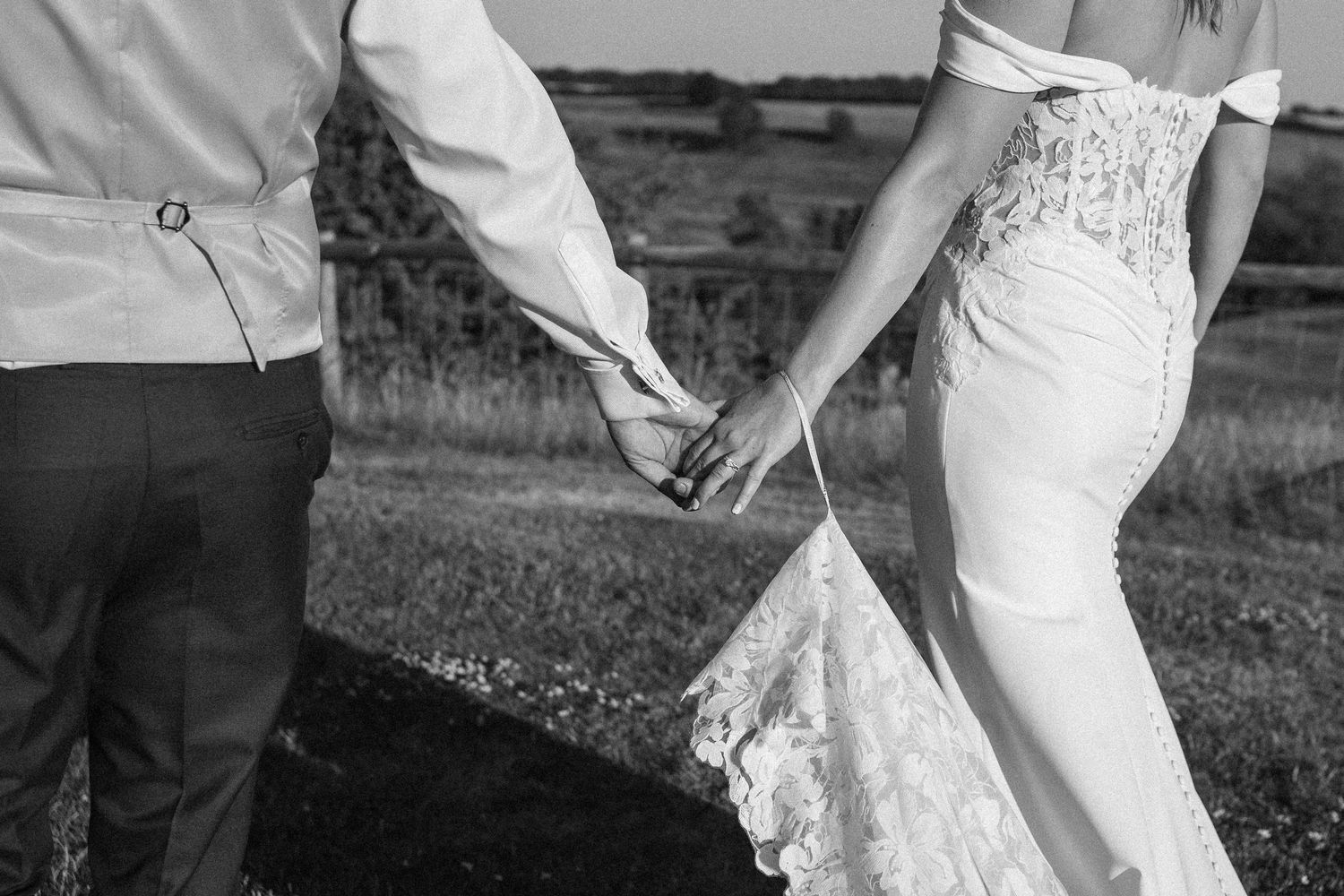 Black and white photo of bride and groom holding hands outdoors, showcasing lace wedding dress train.
