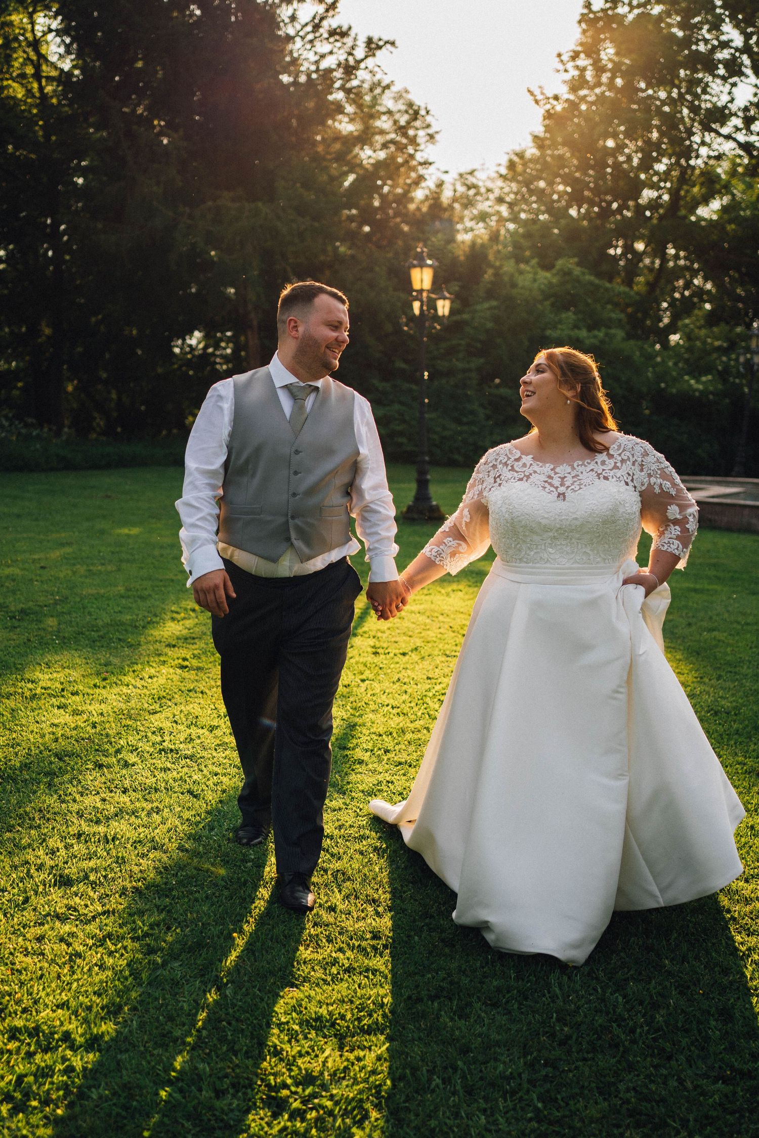 A couple walks hand in hand across a sunlit lawn during golden hour at their outdoor wedding celebration.