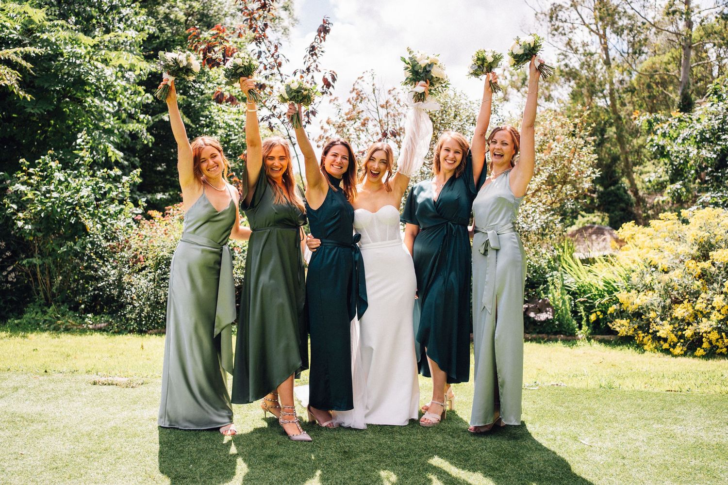 A wedding party poses together outdoors in a garden setting wearing sage green and white dresses on a sunny day.