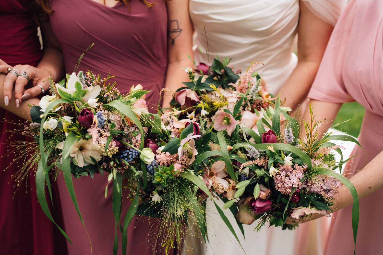 Beautiful wedding bouquets with pink and burgundy flowers mixed with greenery held by bridesmaids in different colored dresses.