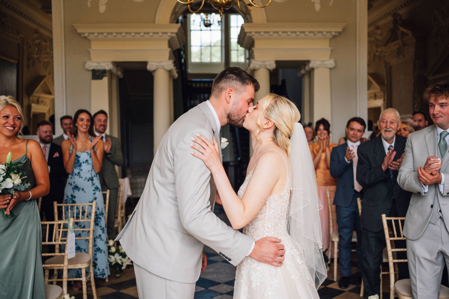 A romantic first kiss moment during a wedding ceremony while guests applaud in an ornate indoor venue.