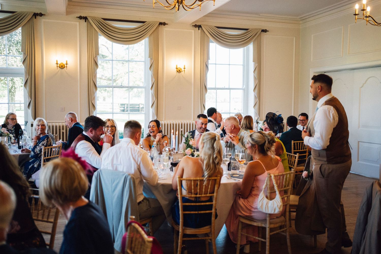 A wedding ceremony takes place in an elegant white room with large windows and wooden chairs.