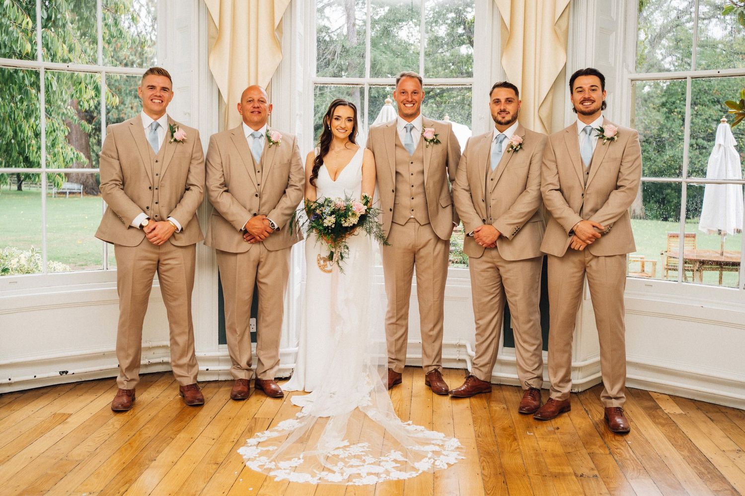 Wedding party in tan suits and blue dresses poses together in a bright sunlit venue.