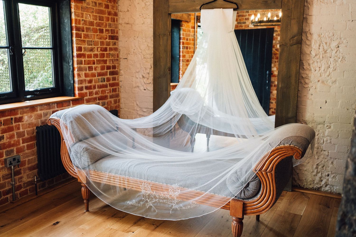Flowing white tulle veil drapes elegantly over a vintage wooden chaise lounge against exposed brick walls.