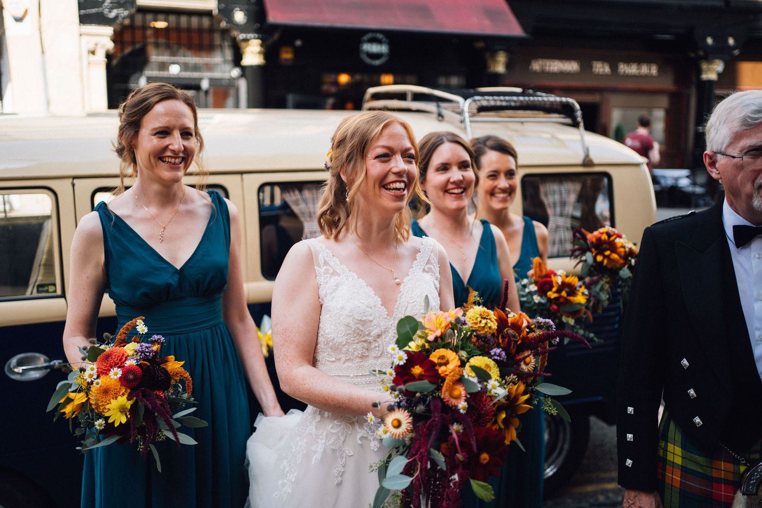 Wedding party members in teal dresses and suits pose together with vibrant orange and red bouquets.