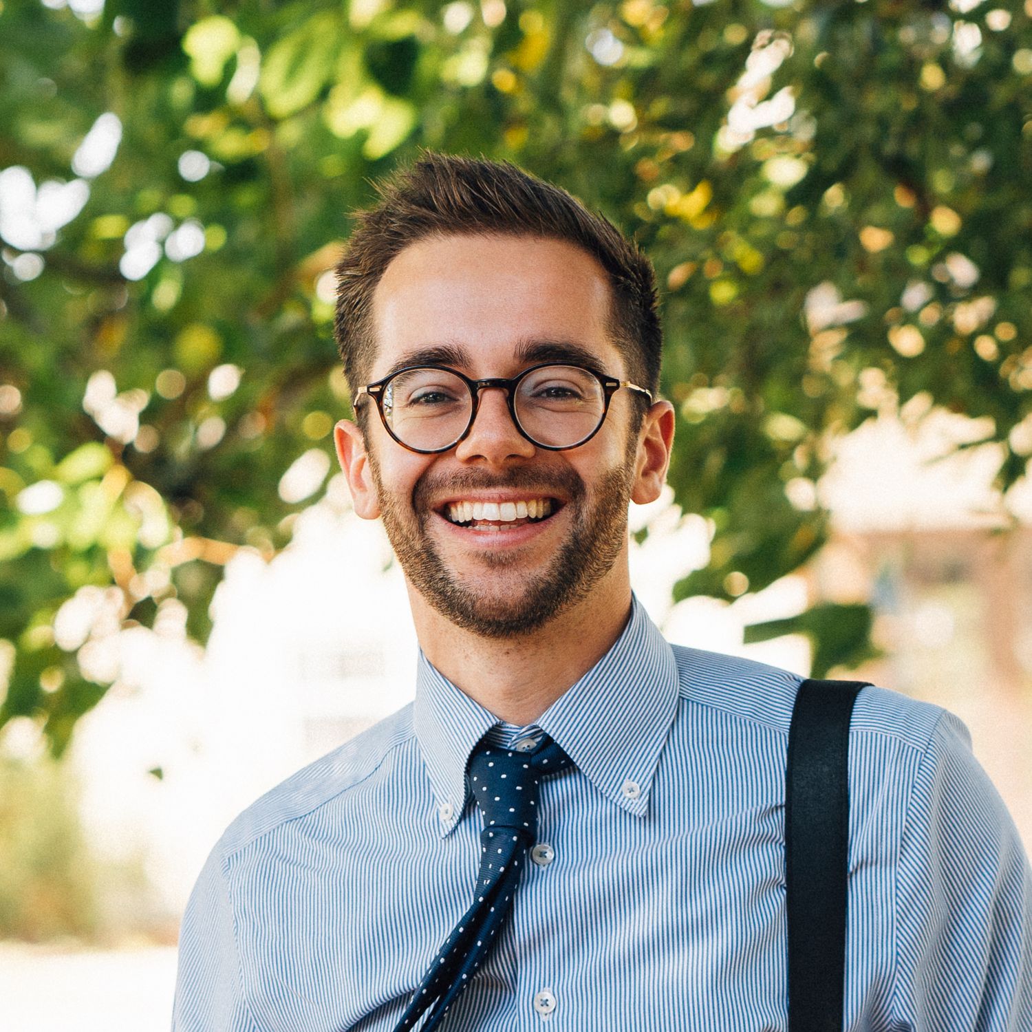 Stylish groom in light blue shirt, navy tie and suspenders smiling outdoors.