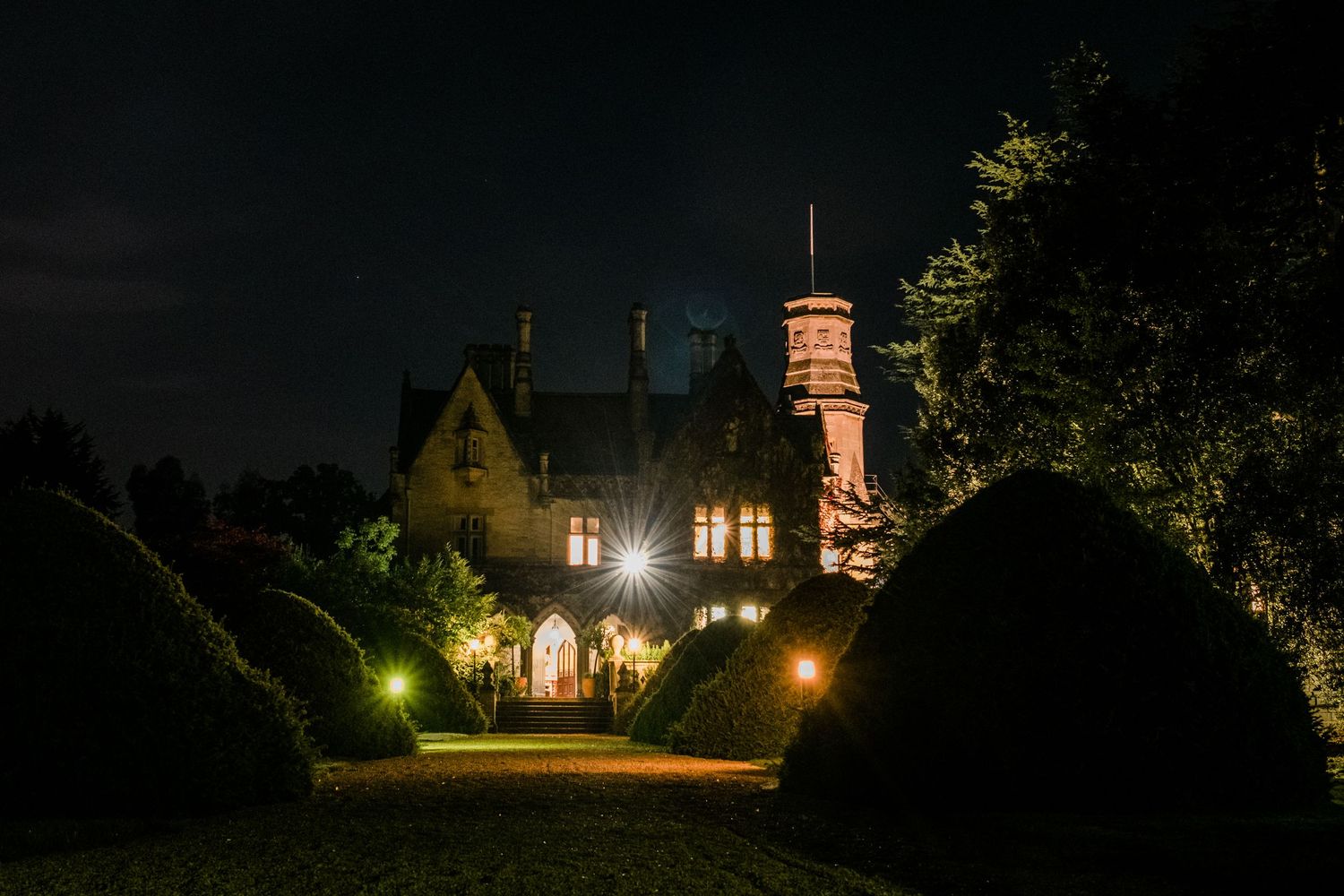Historic stone building illuminated at night with warm glowing windows and landscaping.