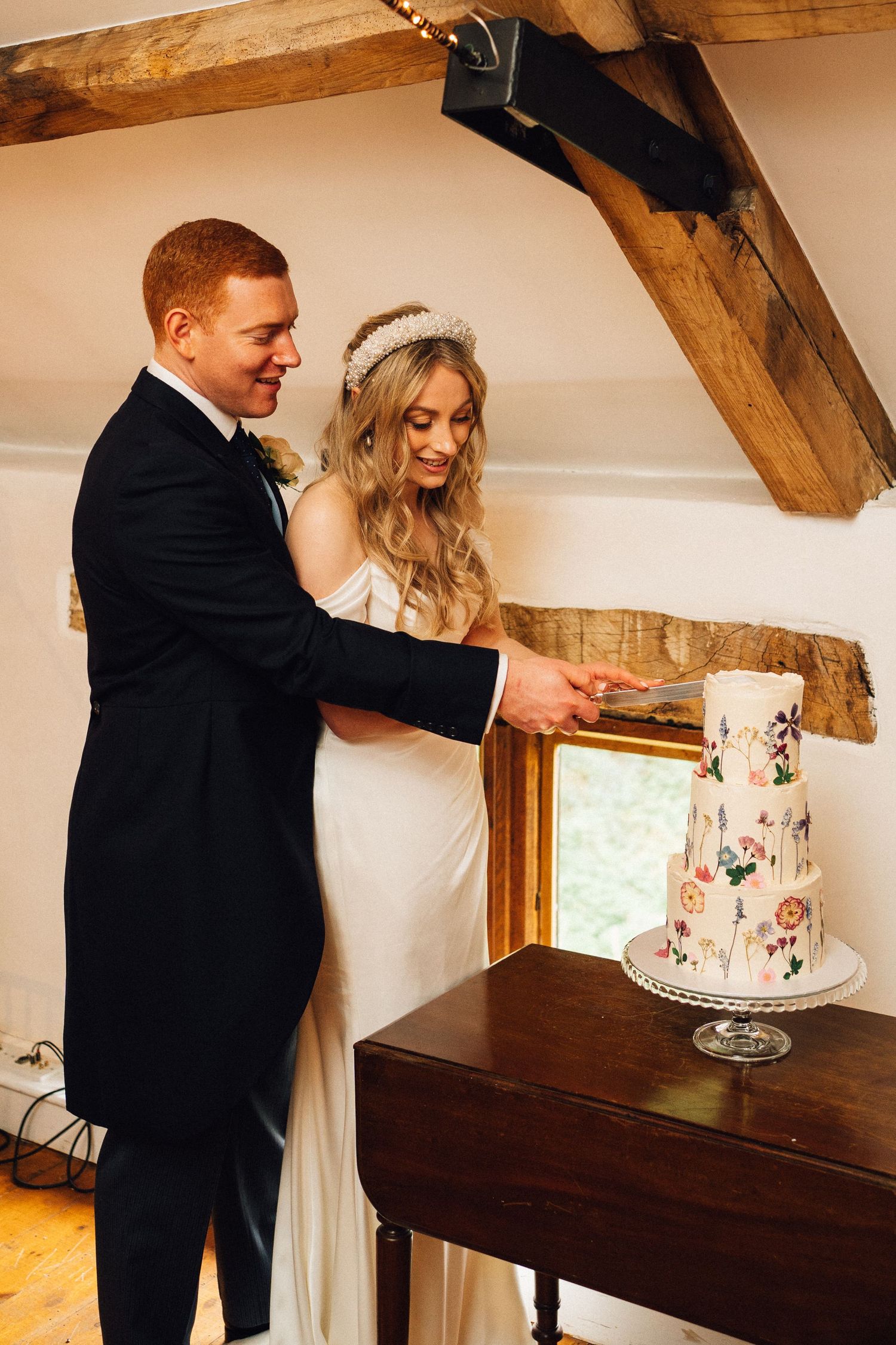 Couple cutting their floral-decorated wedding cake together at a rustic venue.