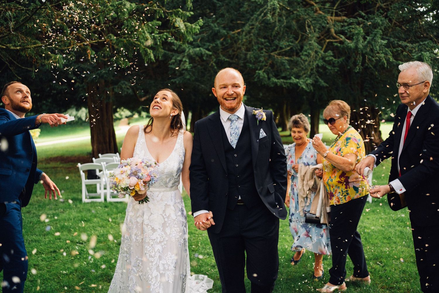 Wedding guests celebrate by throwing confetti on a sunny outdoor lawn.