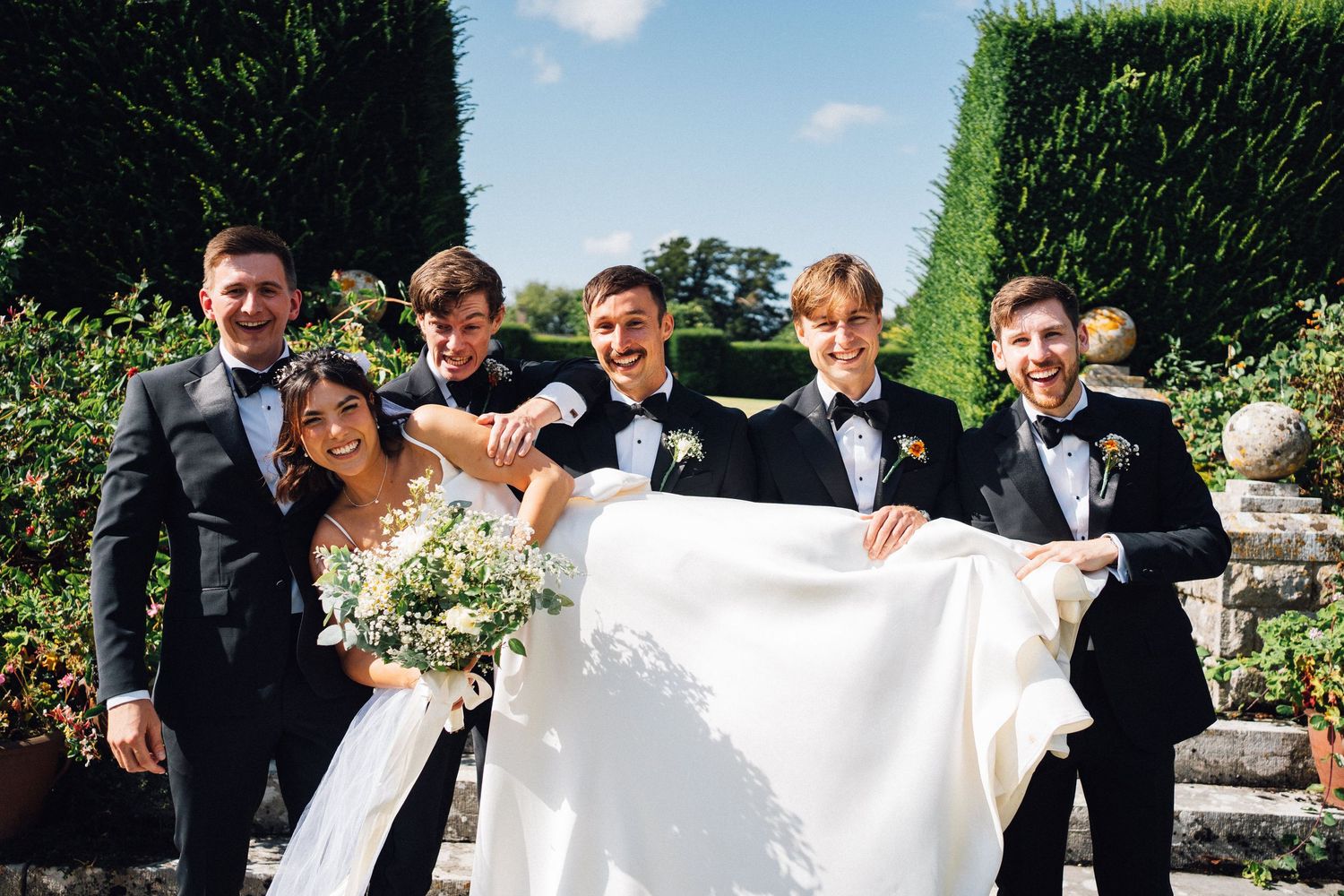 Wedding party creates playful moment with dress train in garden setting.
