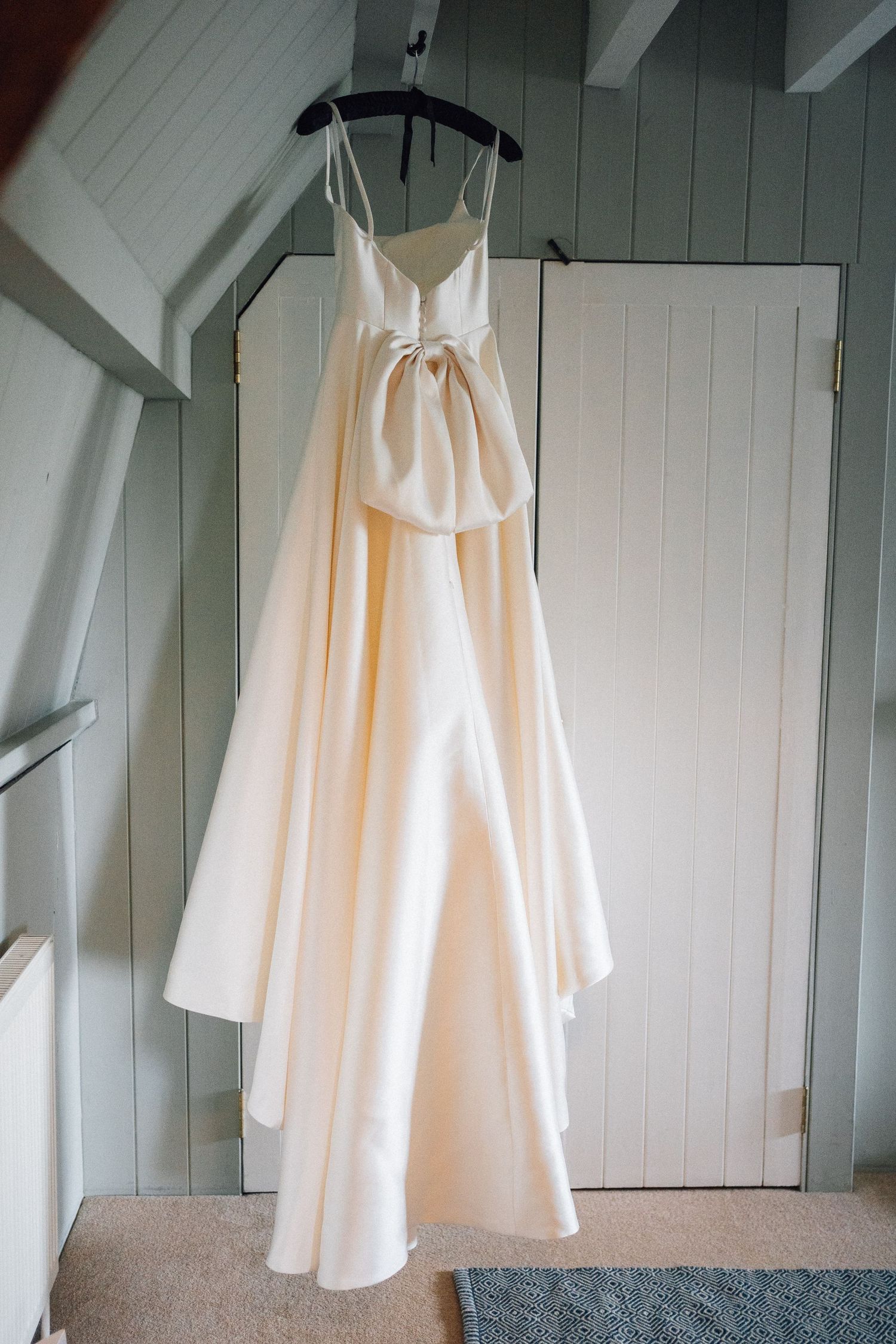 White wedding dress hanging against pale grey wooden paneled wall in natural light.