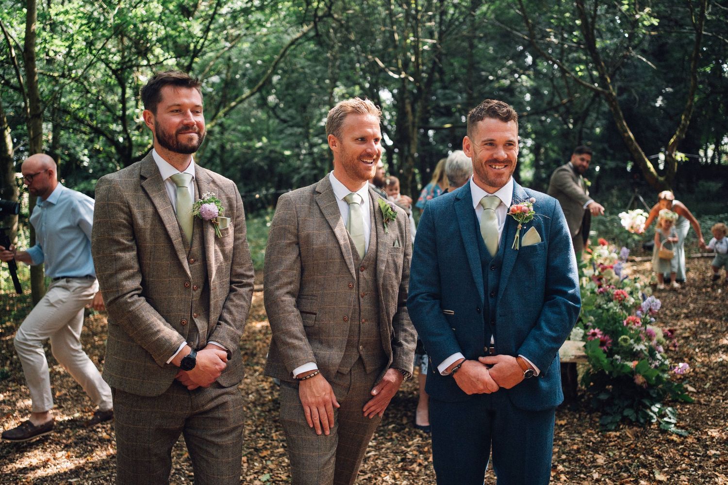 Groomsmen in brown suits pose together under trees at an outdoor wedding.