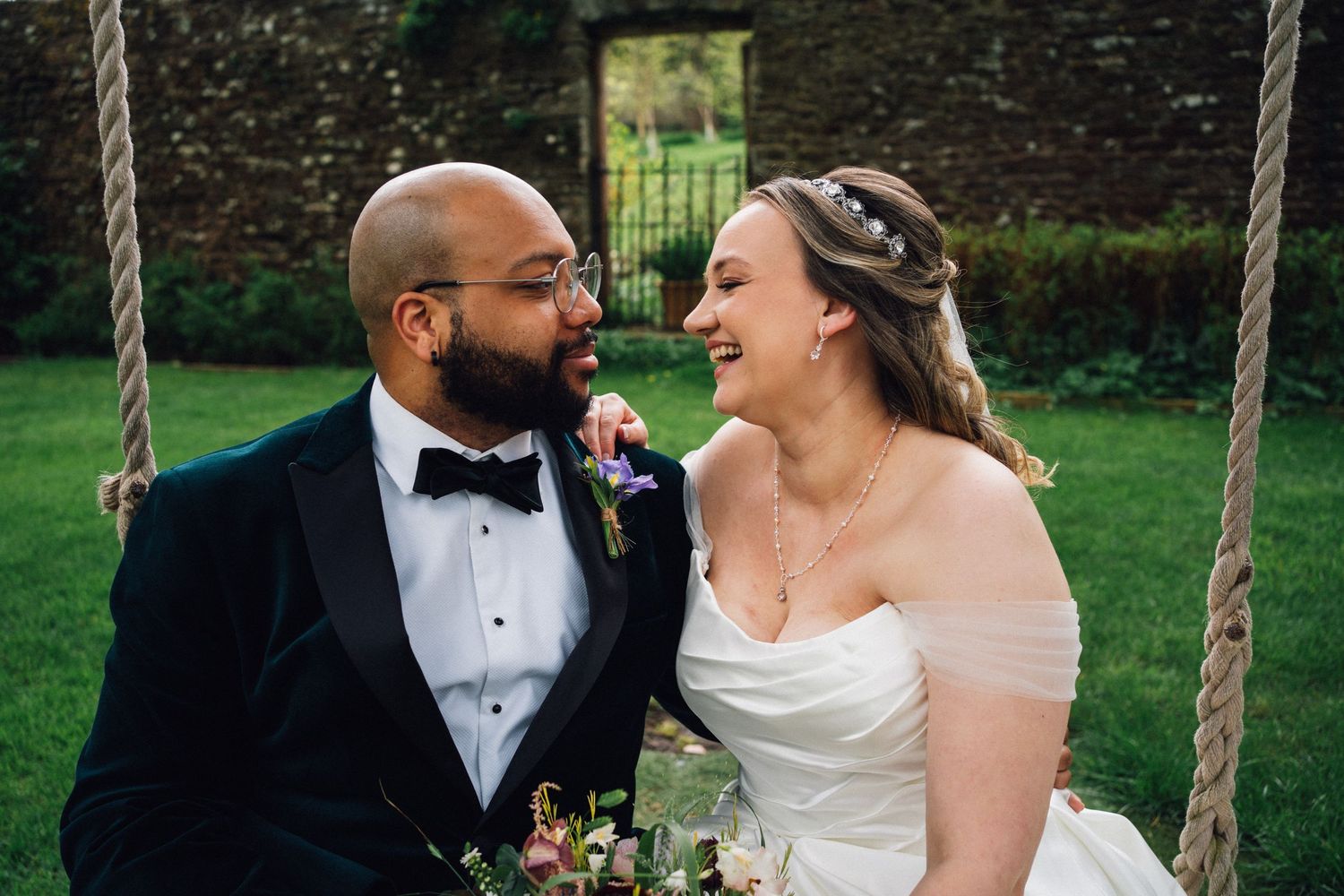 A joyful wedding couple shares a candid laughing moment while sitting on a garden swing.