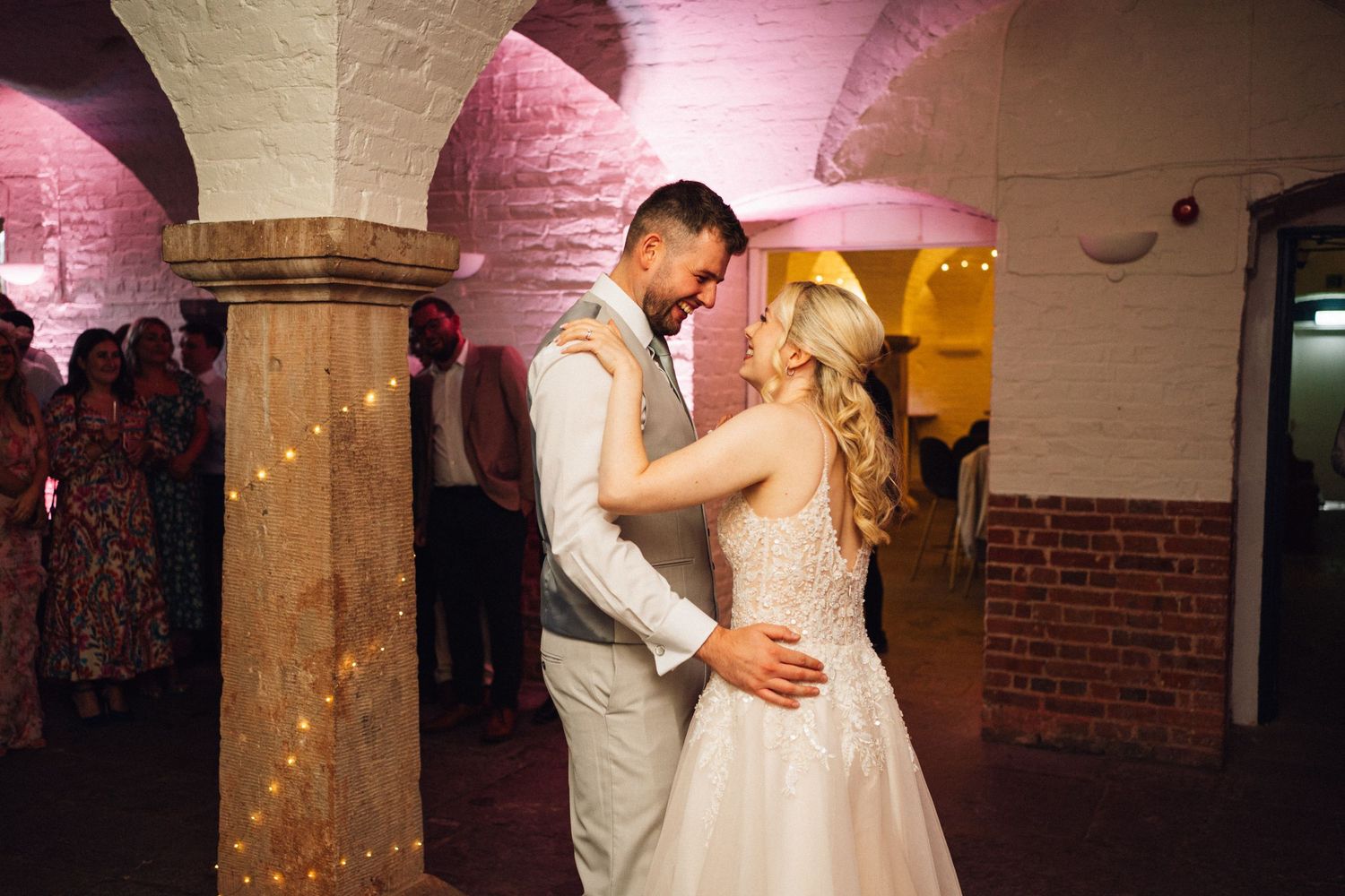 A couple shares their first dance in a romantic brick venue with pink uplighting and stone columns.