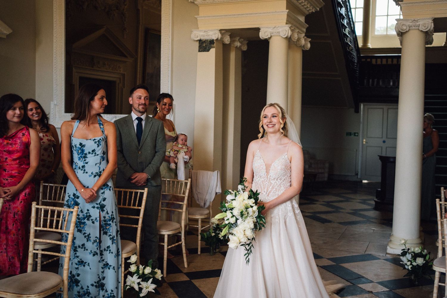 Wedding ceremony scene with guests seated in elegant columned venue with floral arrangements.