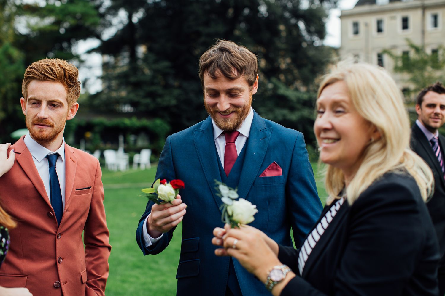 Wedding guests in formal attire share a lighthearted moment while holding flowers in an outdoor garden setting.