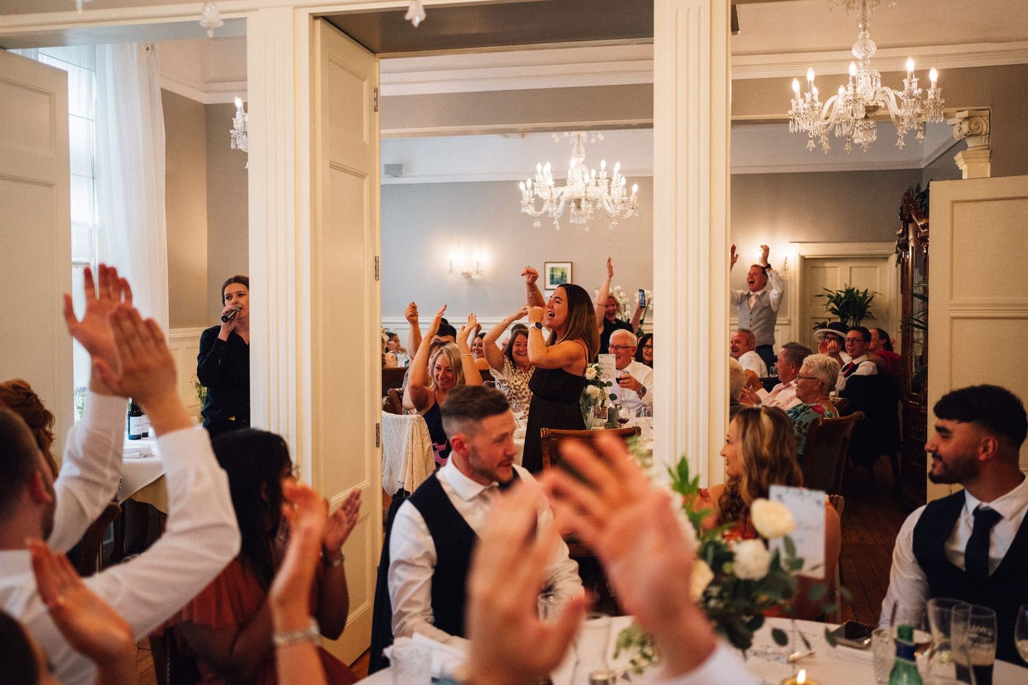 Wedding reception guests raise their hands in celebration under elegant crystal chandeliers in an indoor venue.