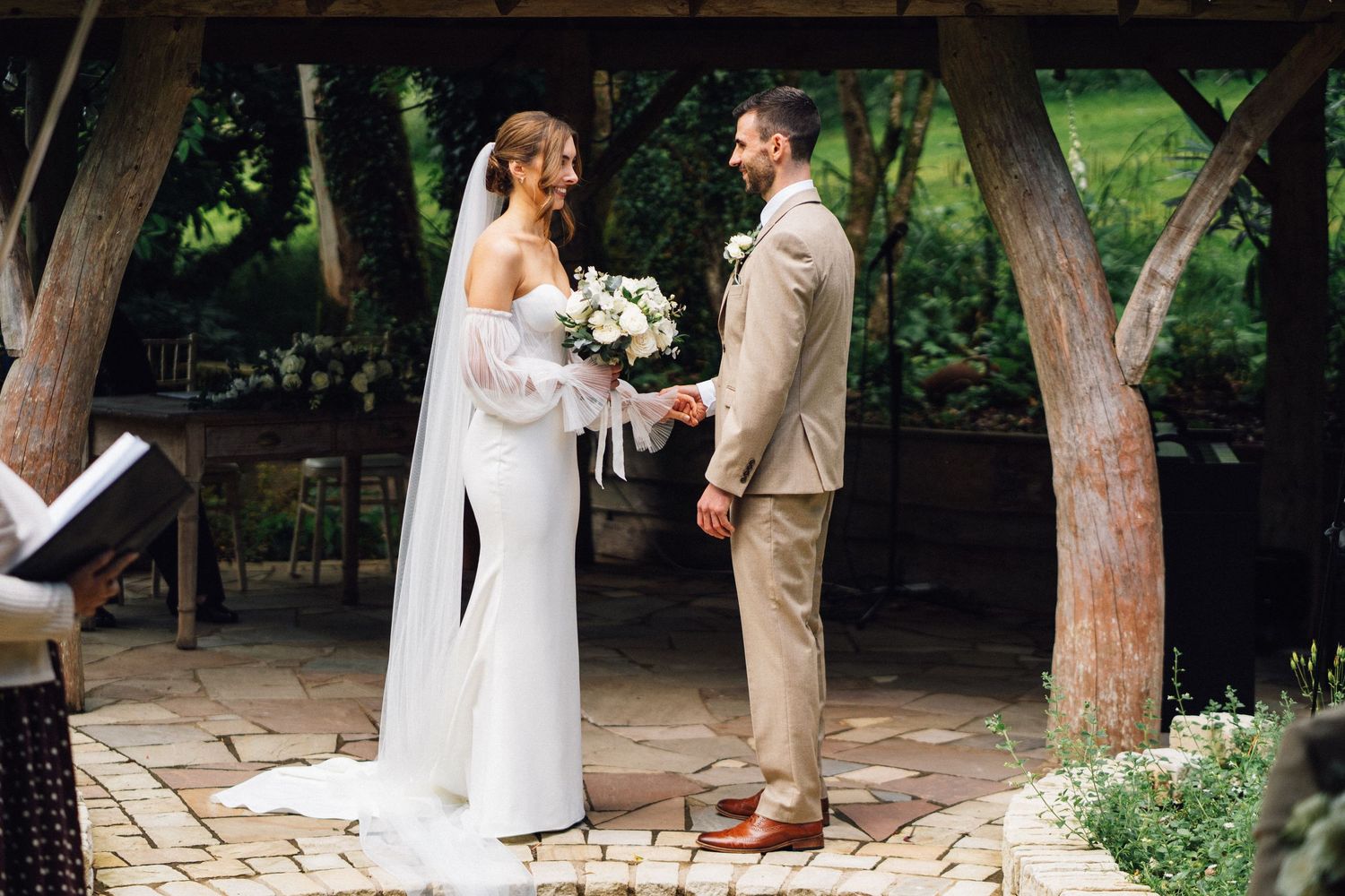 A couple exchanges vows during an intimate outdoor wedding ceremony beneath tall trees.