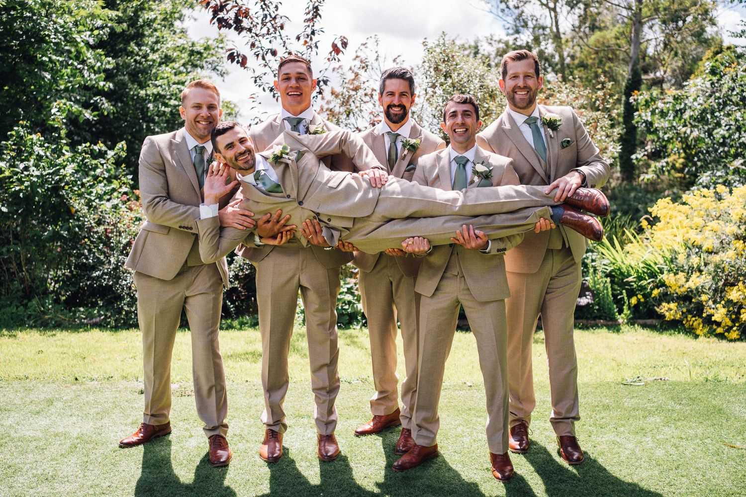 Groomsmen in tan suits playfully carry a person horizontally for a fun wedding photo in a garden setting.