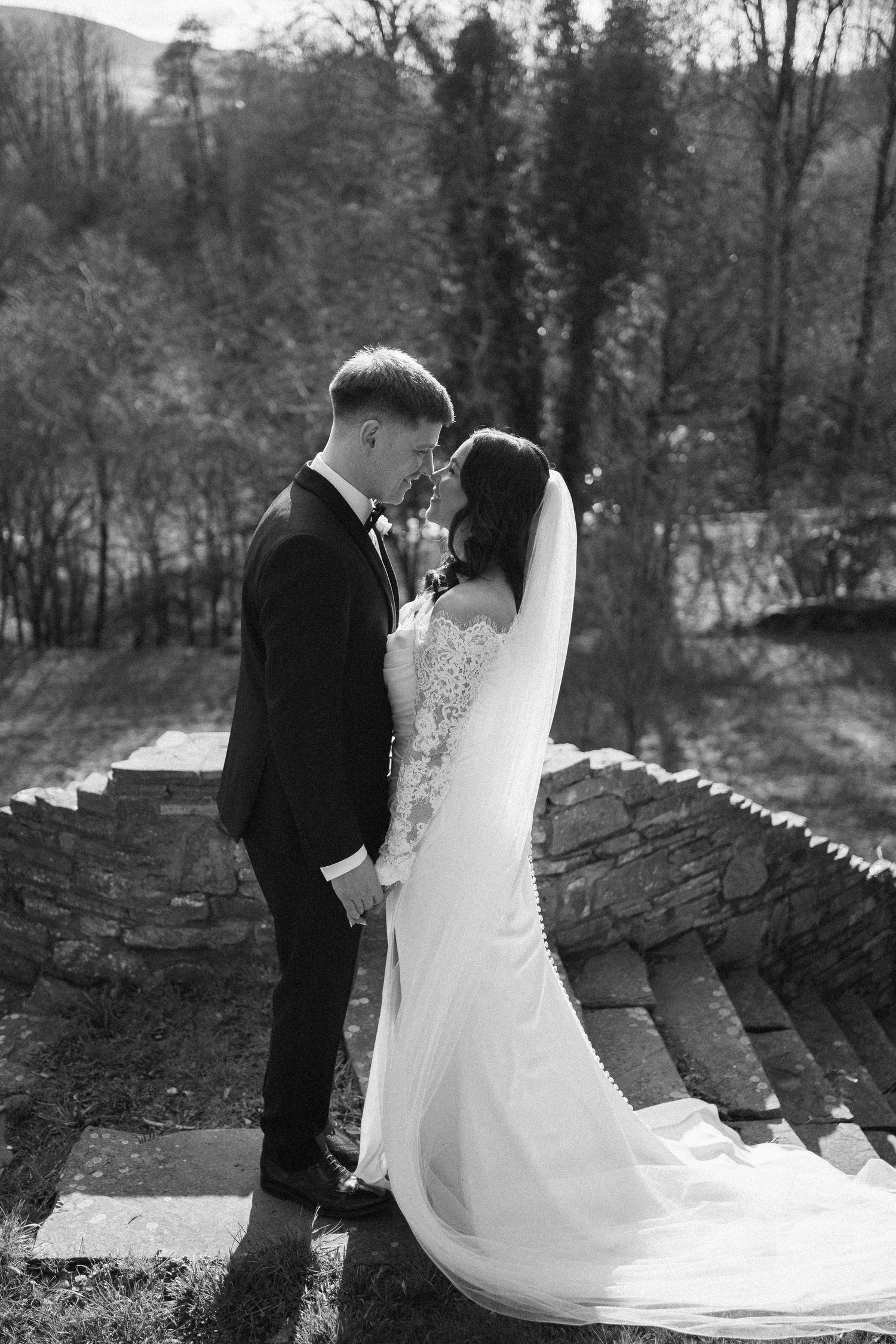 Black and white wedding photo of bride and groom holding hands, standing on stone steps with trees in background.