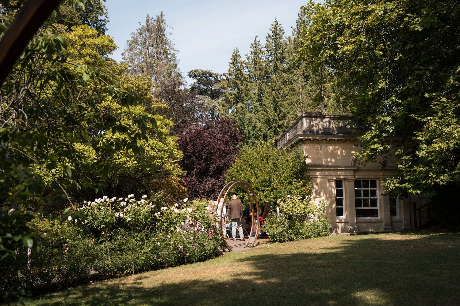 Lush garden with flowering borders leading to a charming stone building surrounded by tall trees on a sunny day.