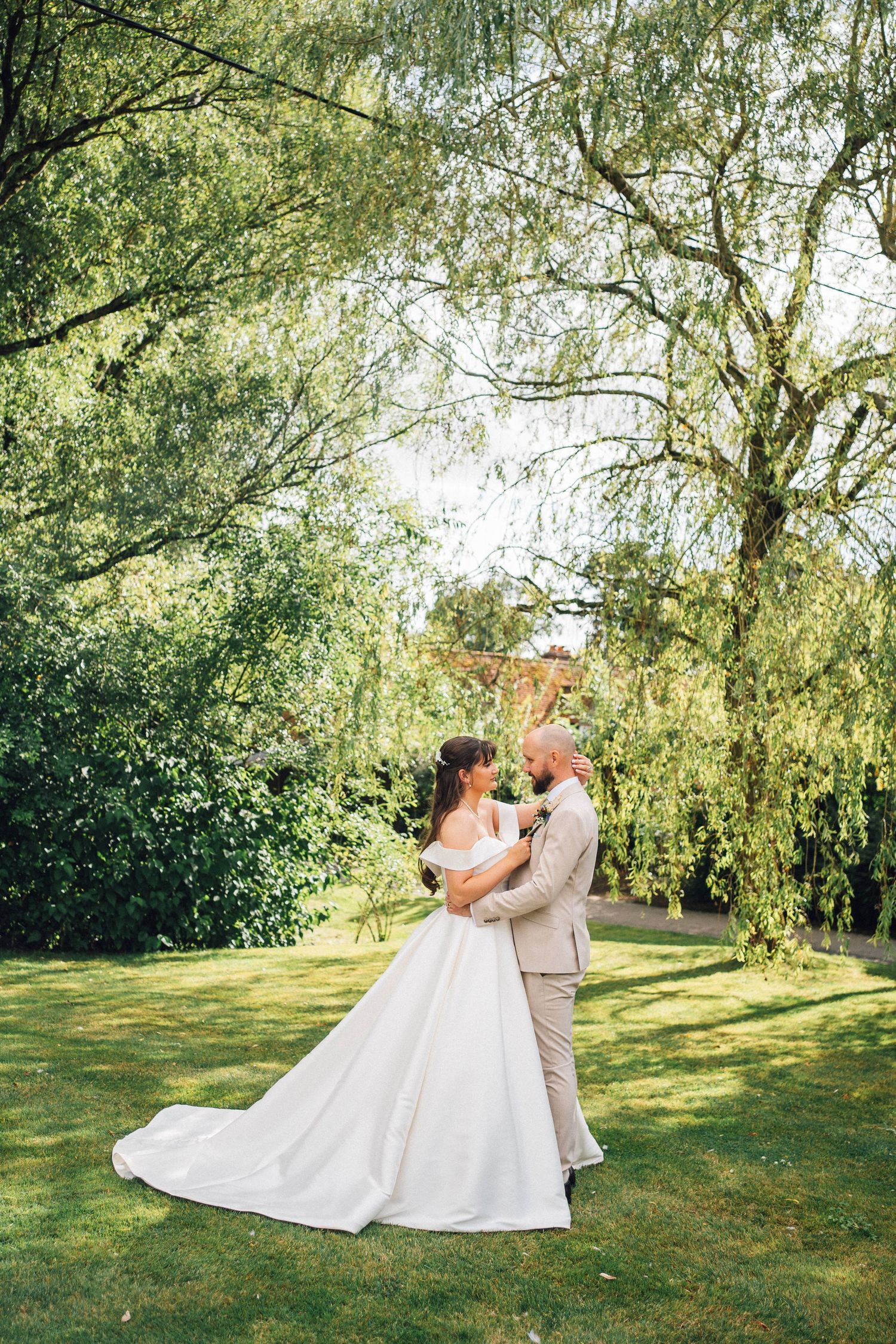 A bride in a white ball gown and groom in a beige suit share a romantic moment in a lush green garden.