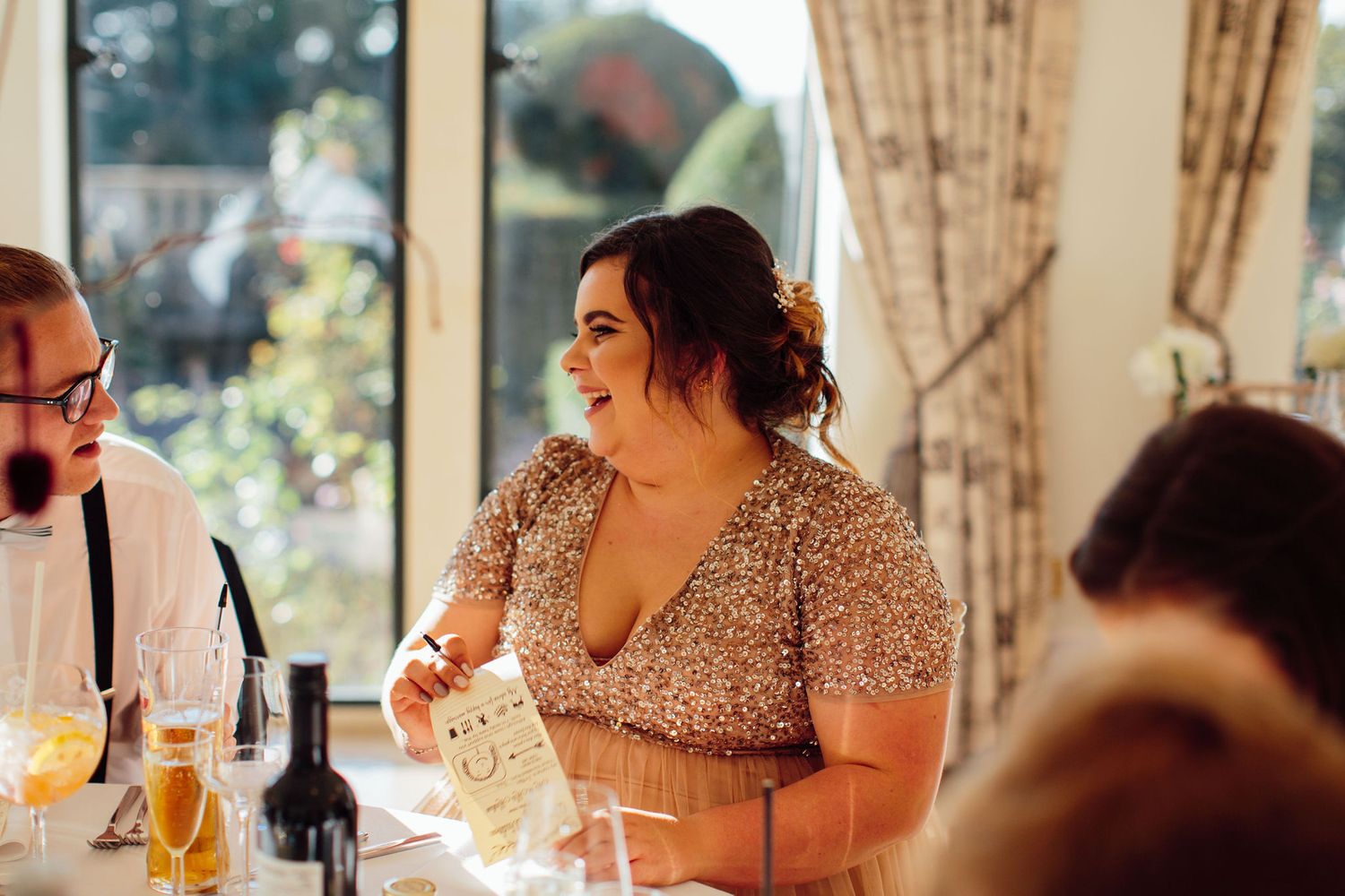 Wedding reception guests seated at decorated tables in an elegant venue with natural lighting.