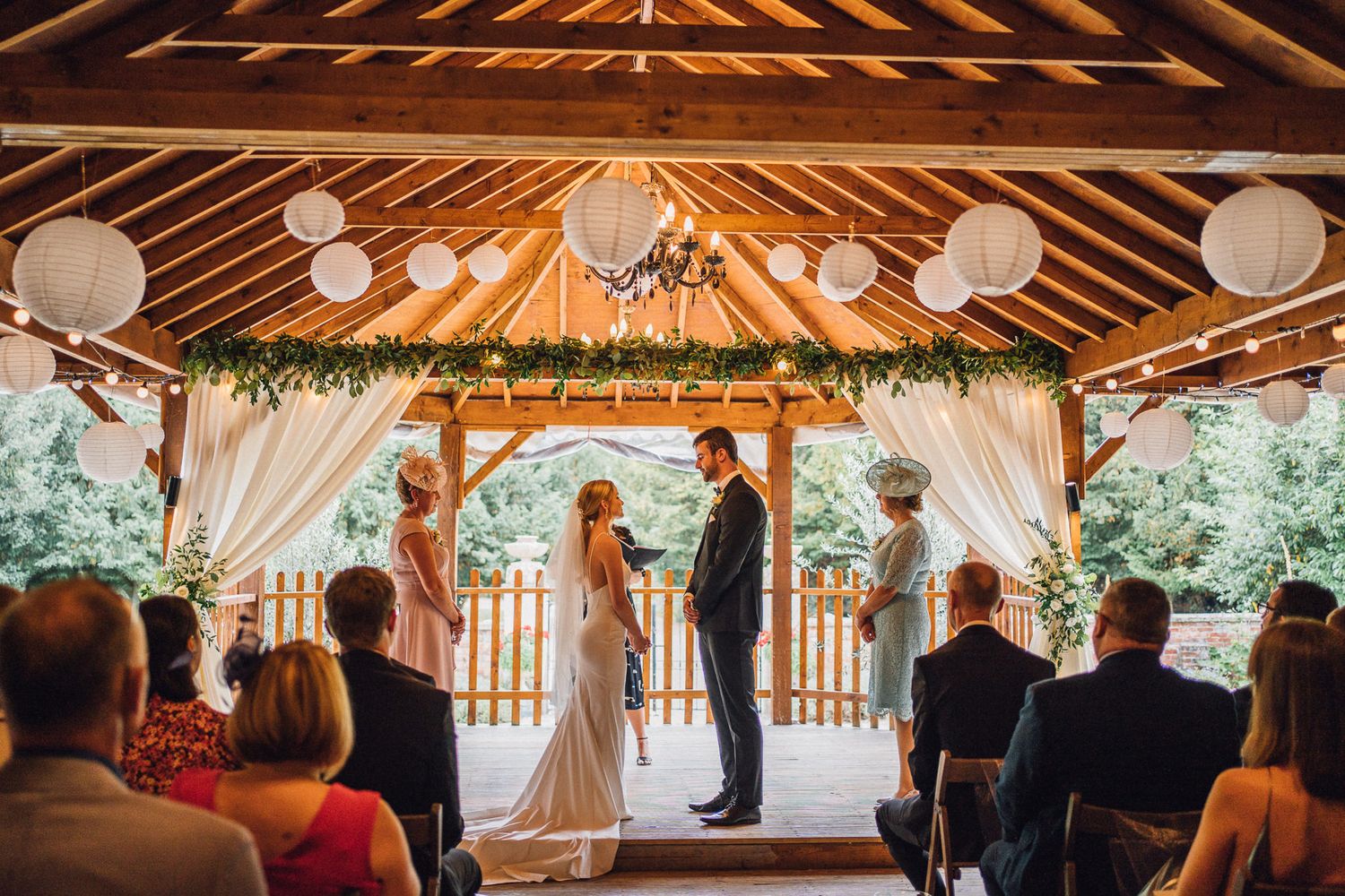 Guests seated in a rustic barn venue watching a wedding ceremony under wooden beams and white decorations.