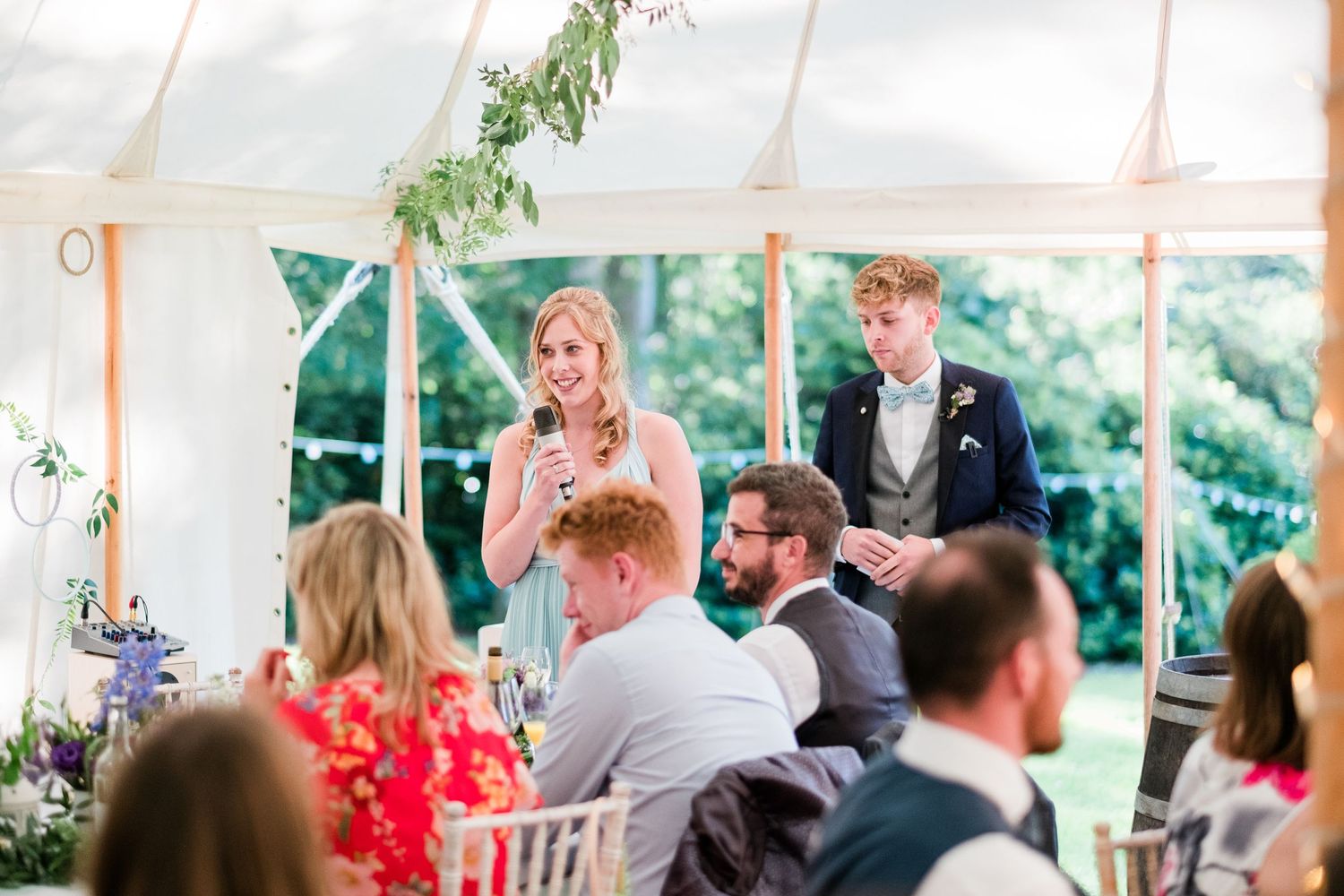 Guests seated at decorated tables during a wedding reception in a white tent with hanging greenery.